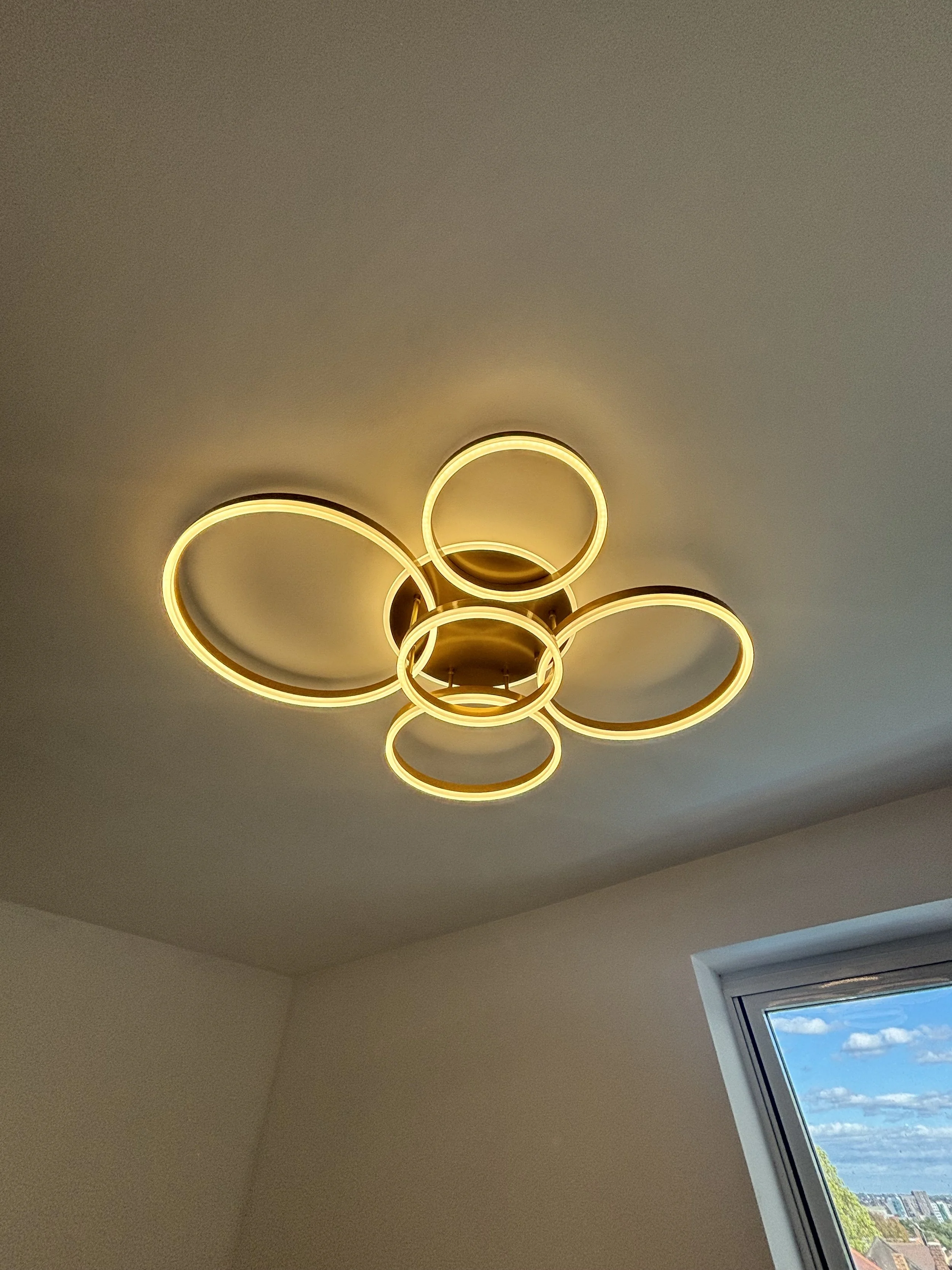 Ceiling light fixture with multiple circular rings emitting warm light, mounted on a beige ceiling in a room with an angled window showing a view of partly cloudy sky and trees outside.