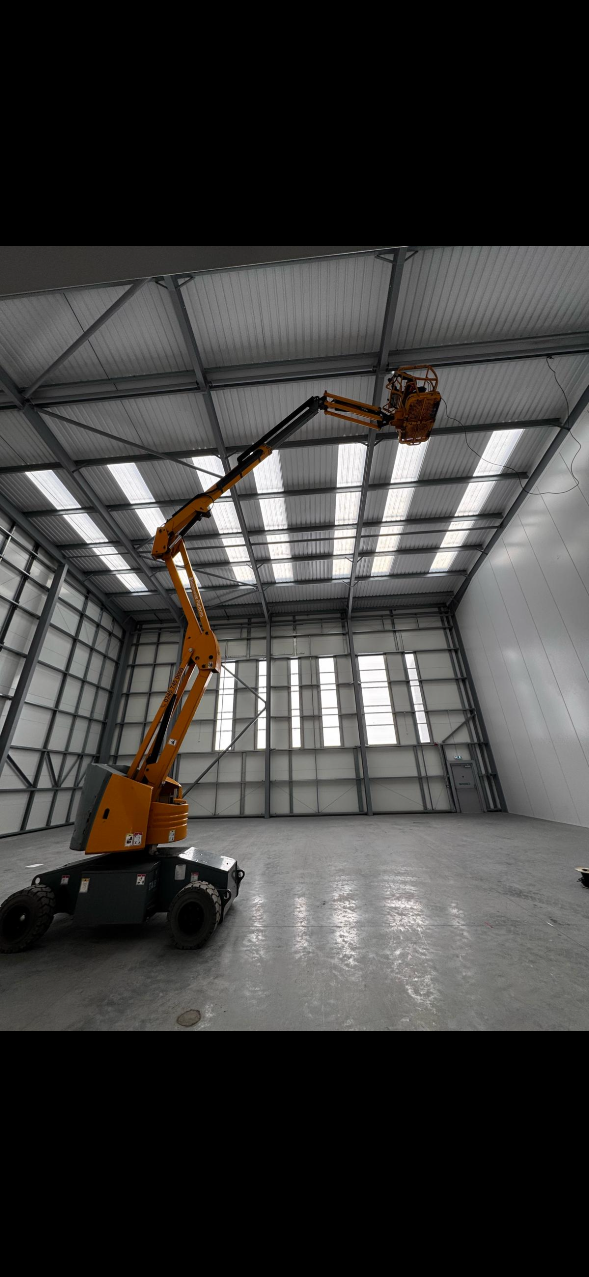 Inside a large industrial warehouse with a yellow cherry picker lift extended towards the ceiling. The warehouse has a metal roof with skylights and large windows on the walls.