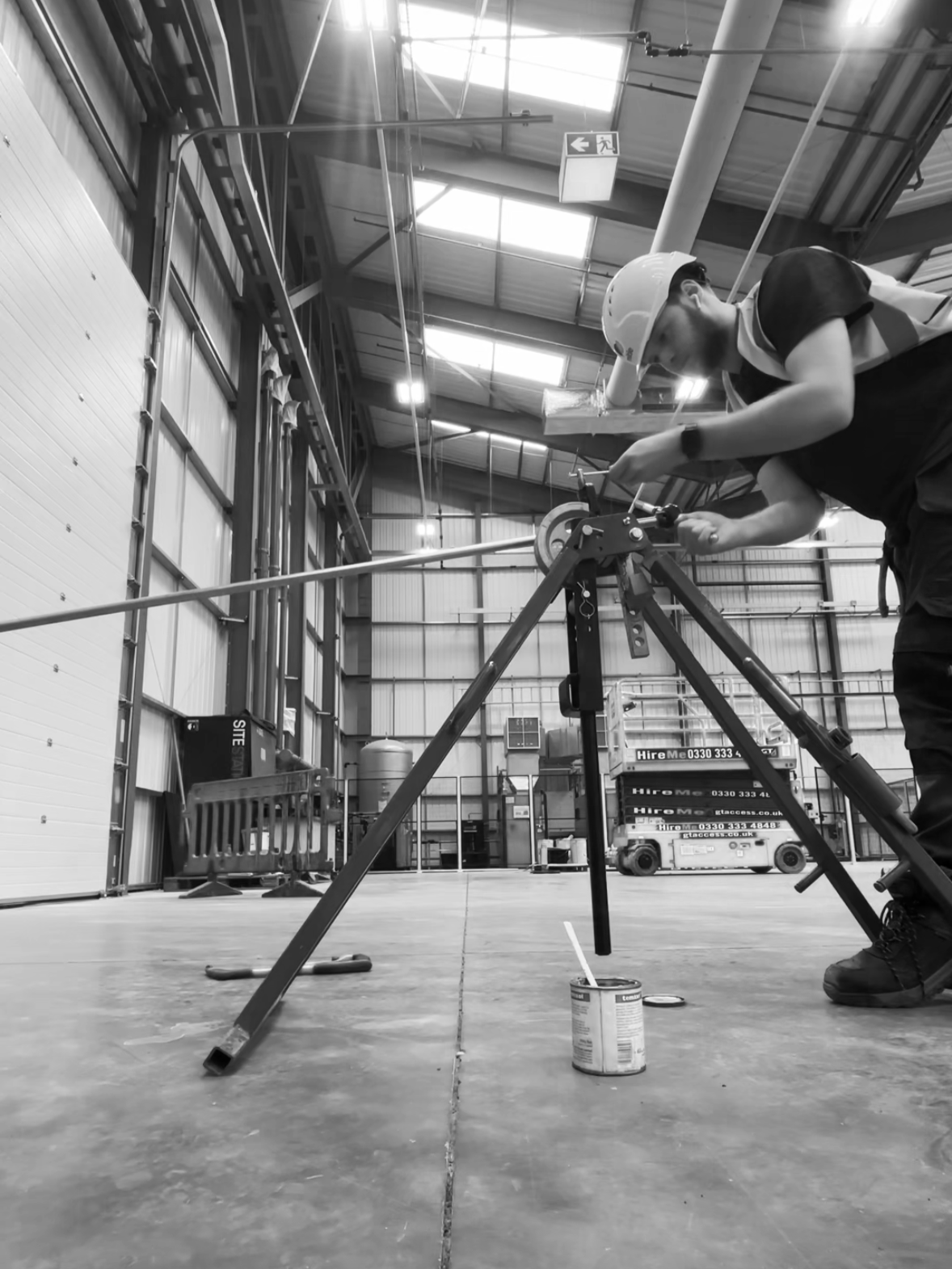 A worker wearing a safety helmet using a theodolite or similar surveying instrument inside a large industrial warehouse.