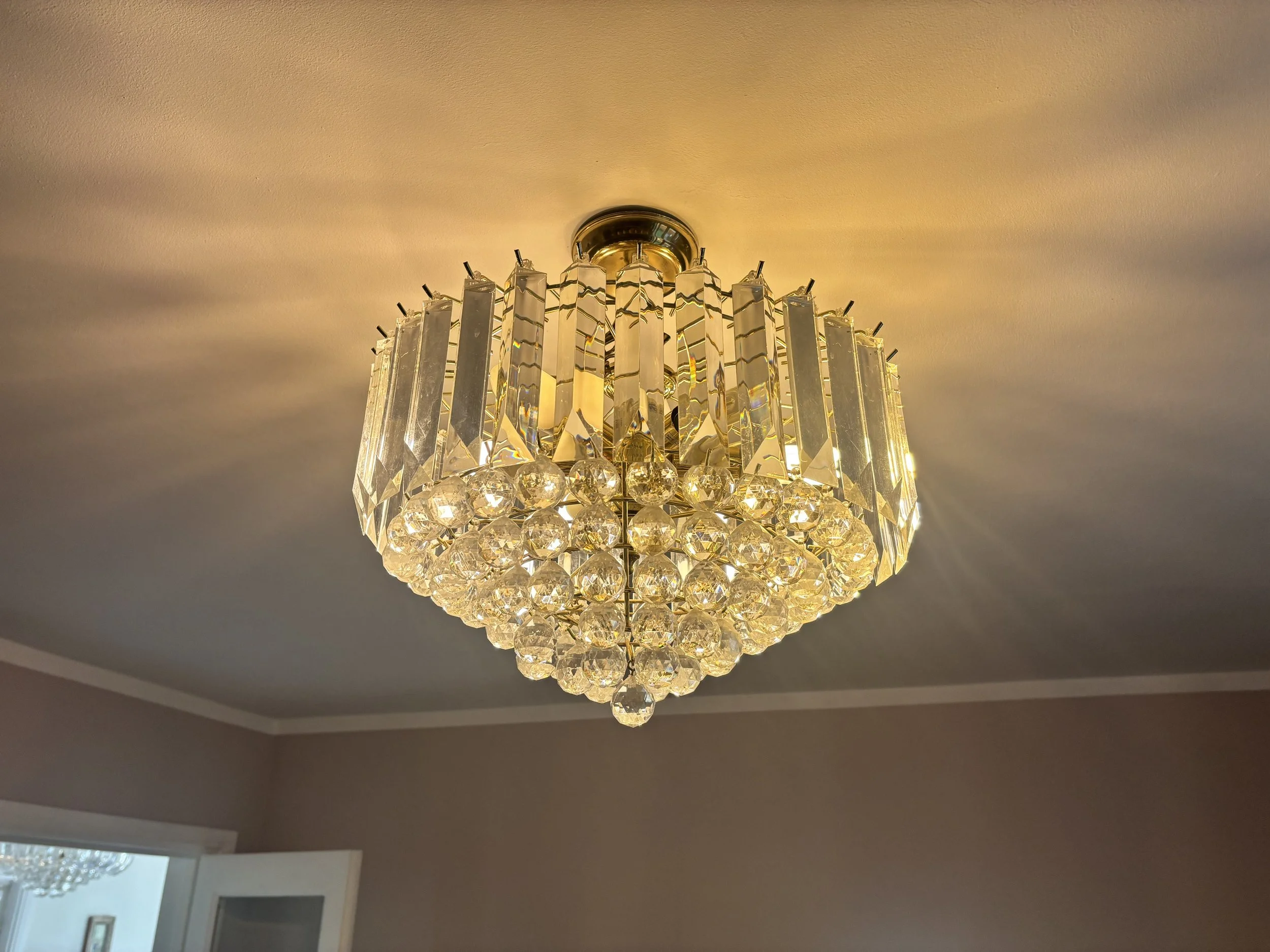 A decorative chandelier hanging from a ceiling, adorned with multiple crystals, with wall and window in the background.