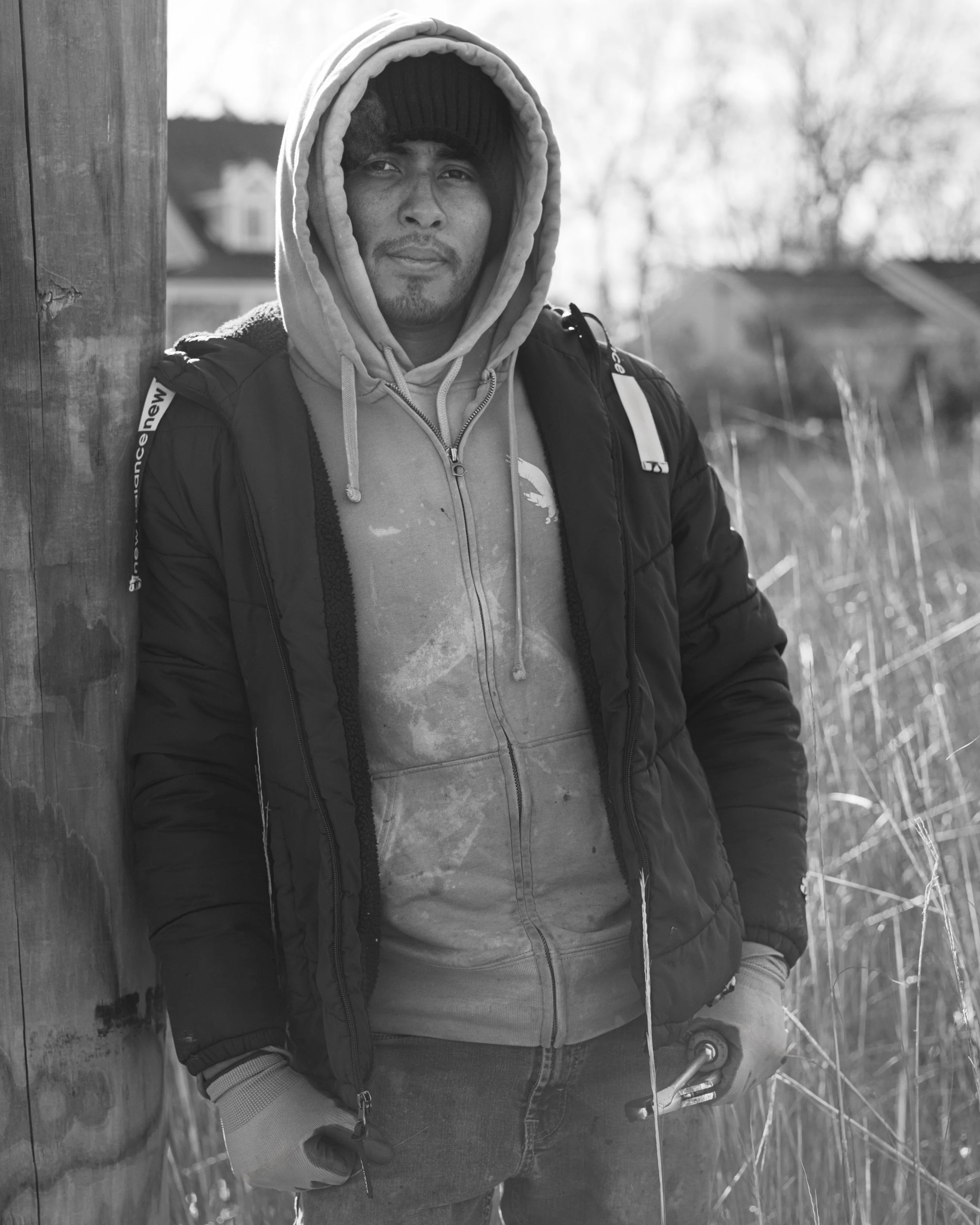 A young man wearing a hoodie, a beanie, and a winter jacket outdoors, leaning against a wooden post, with a blurred background of trees and structures in black and white.
