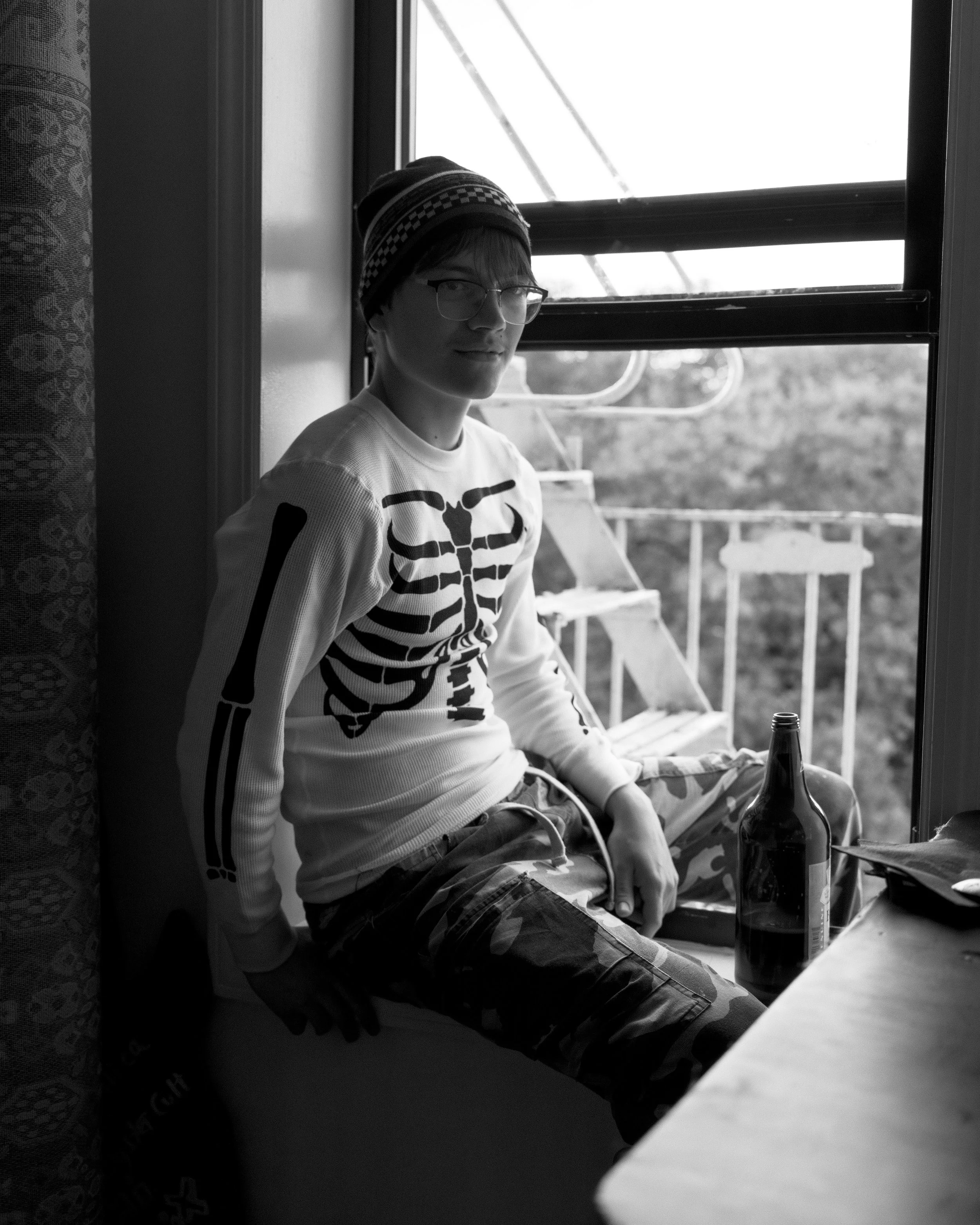 A young man with glasses and a striped winter hat, sitting near a window, holding a smartphone, with beer bottles on a table and a background of trees outside.