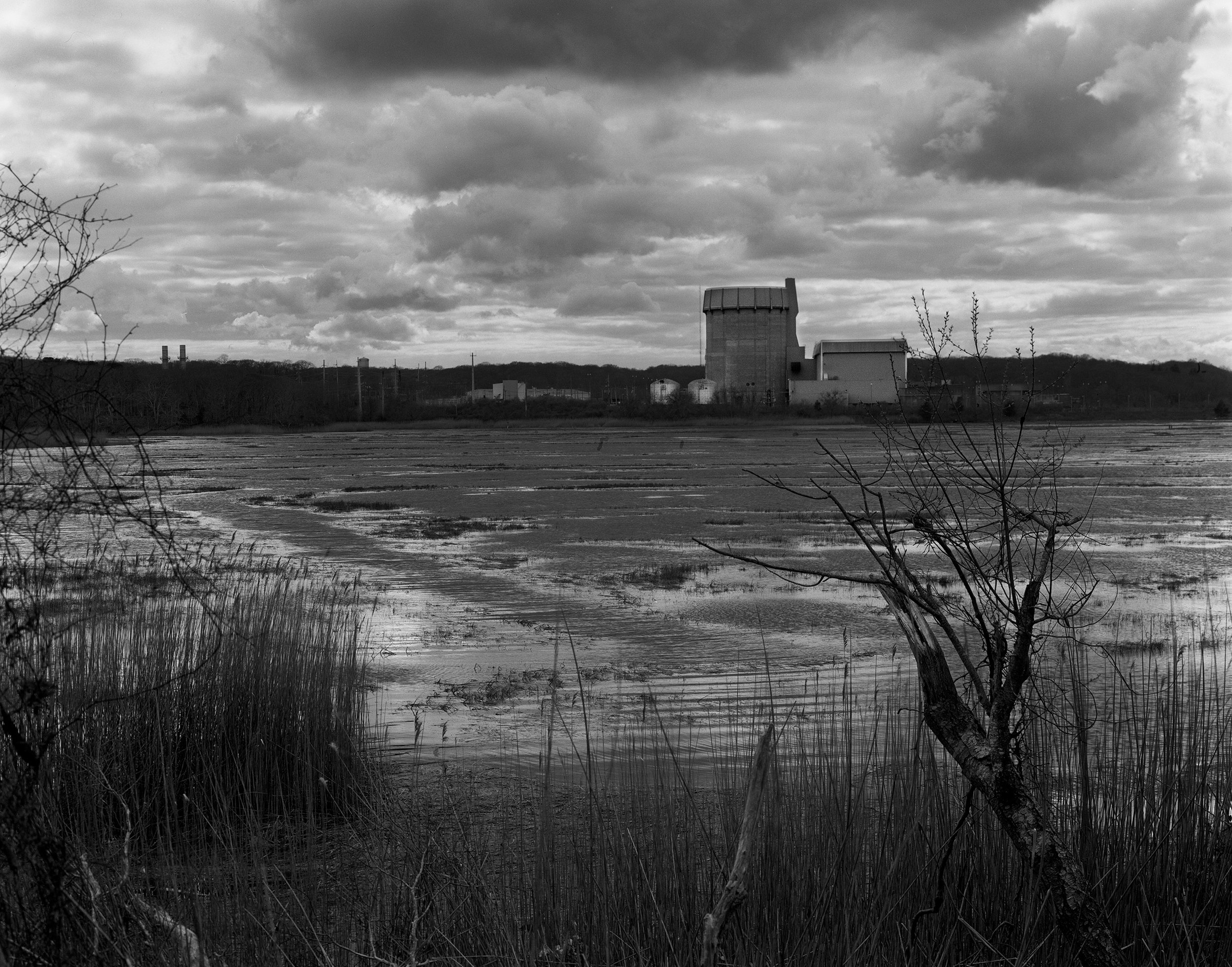 A black and white landscape featuring a body of water with grass in the foreground, a large industrial cooling tower, and a cloudy sky in the background.