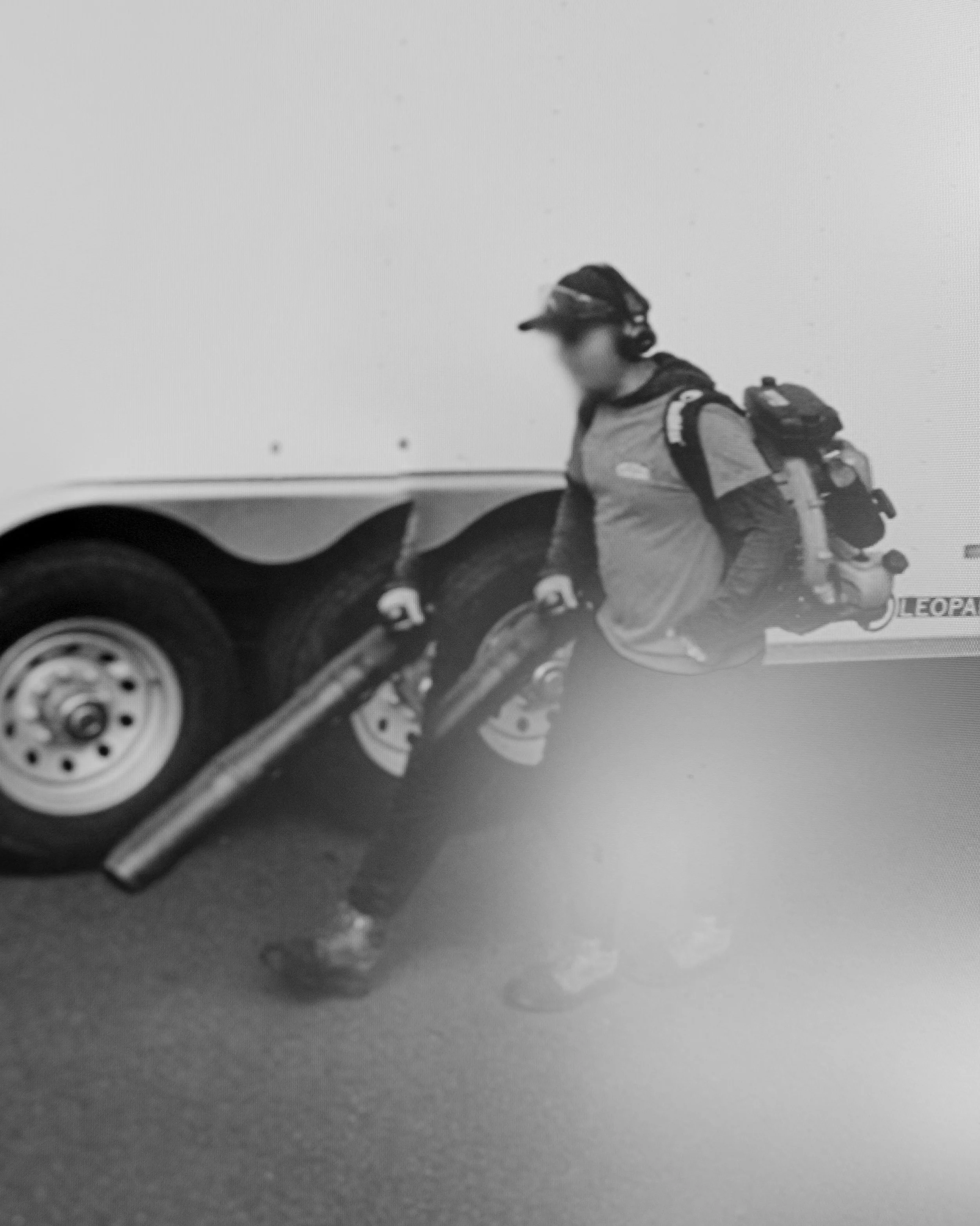 Black and white photo of a person in a motorcycle gear, including a jacket, helmet, and gloves, standing next to a motorcycle. The background features a plain wall and a portion of a vehicle tire.