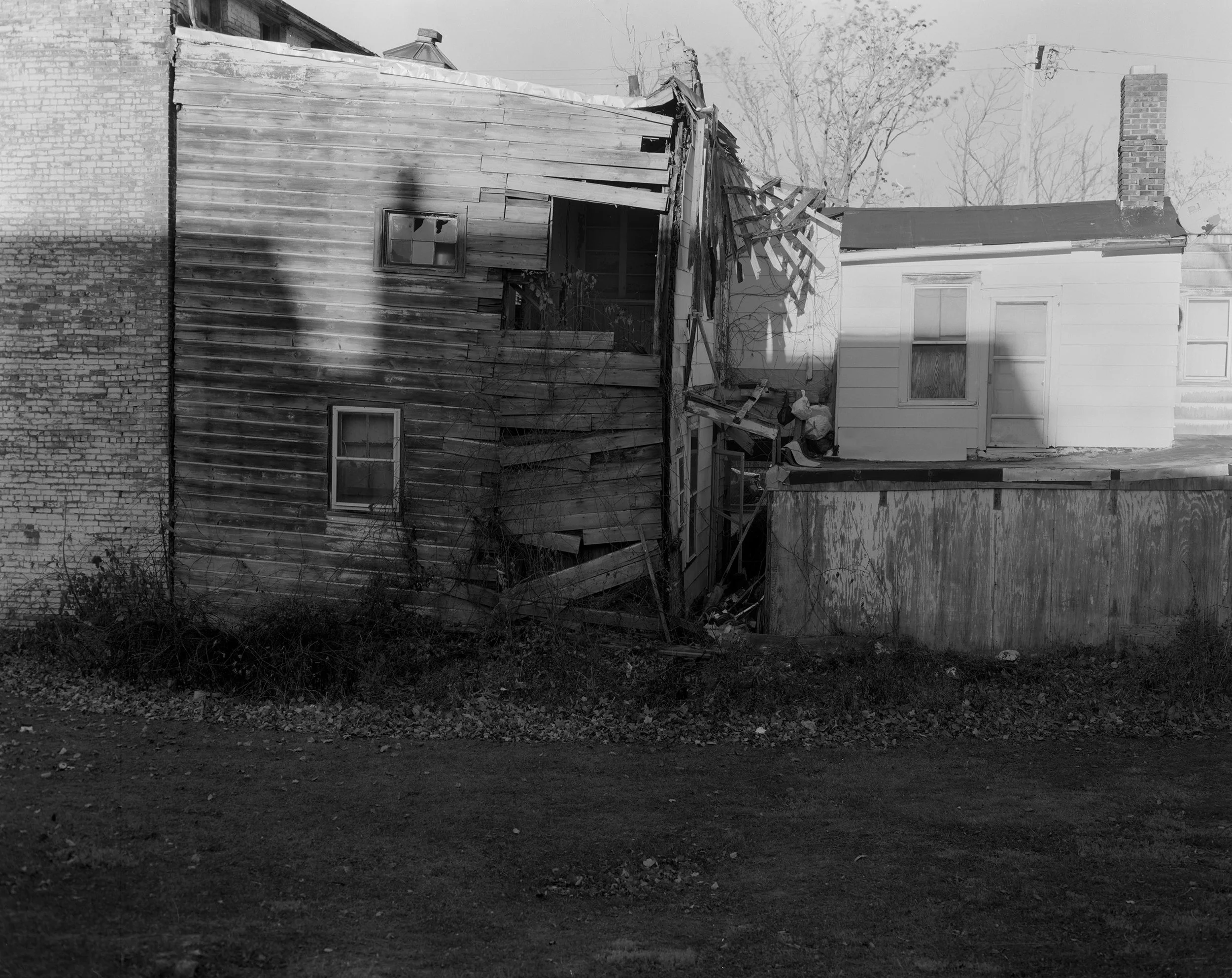 A black and white photograph of a dilapidated house with a damaged roof. The house has a brick chimney and wooden siding, with a new white addition attached. The yard in front is bare dirt, and some trees are visible in the background.