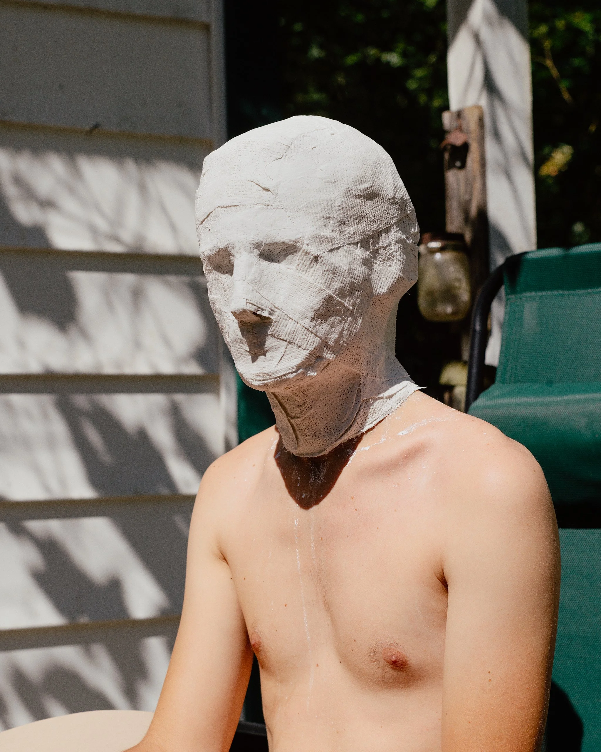 A person is wearing a white plaster sculpture mask and sitting outdoors. The mask covers the entire face and is textured with visible cracks. The person has no shirt on, revealing their shoulders and upper chest. Shadows from nearby objects are cast 