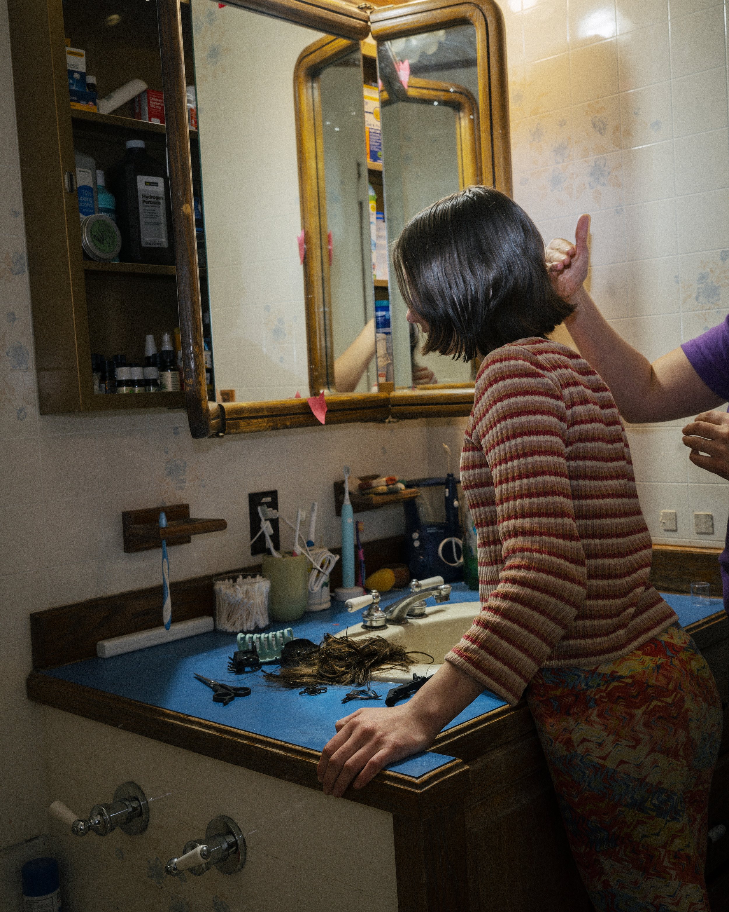 A woman with short dark hair wearing a red and yellow striped shirt getting her hair cut at a bathroom vanity with a blue mat, with hairdressing tools on the counter. A hand holding scissors is seen trimming her hair.