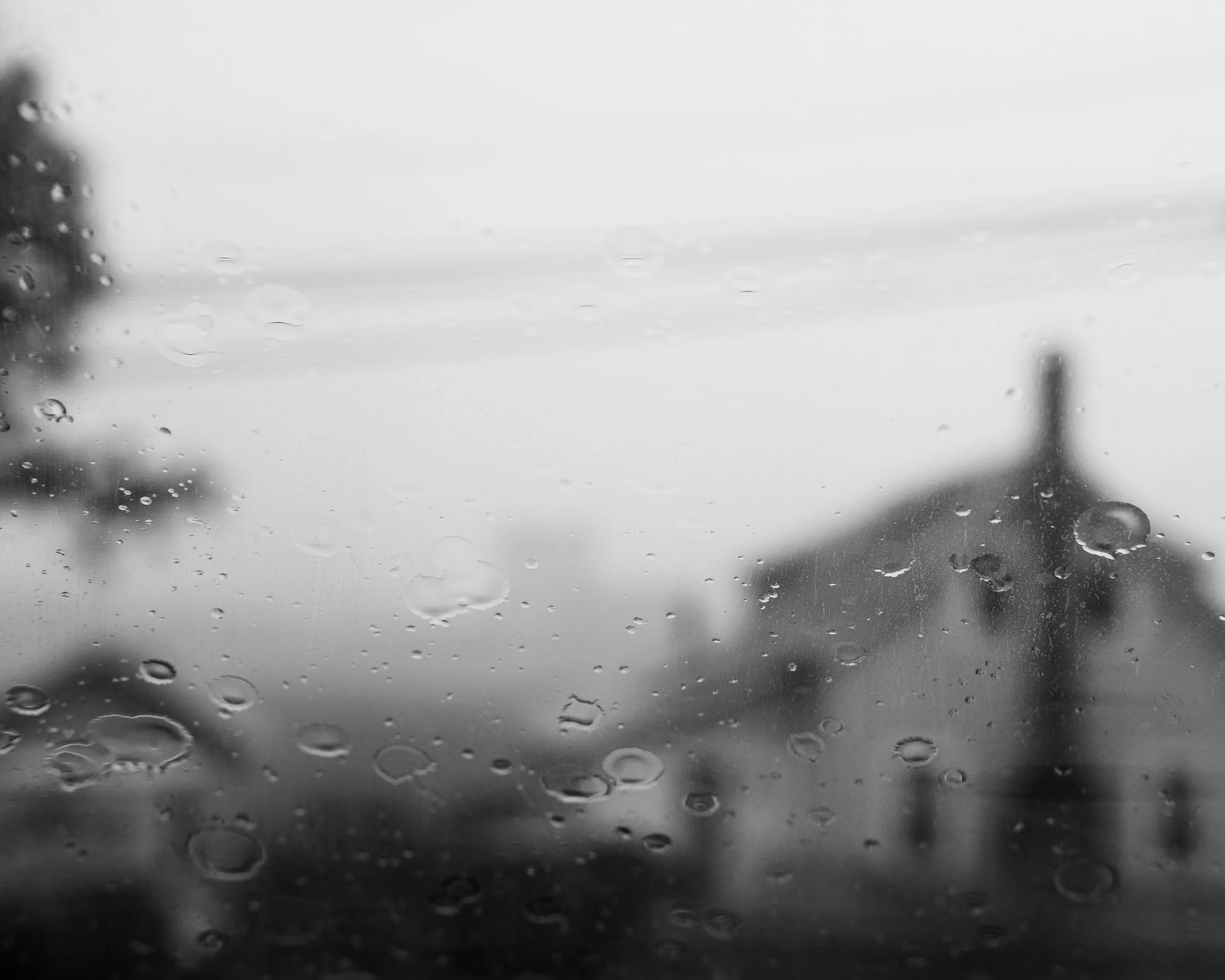 Rain droplets on a glass window with a blurry view of a cloudy sky in the background.