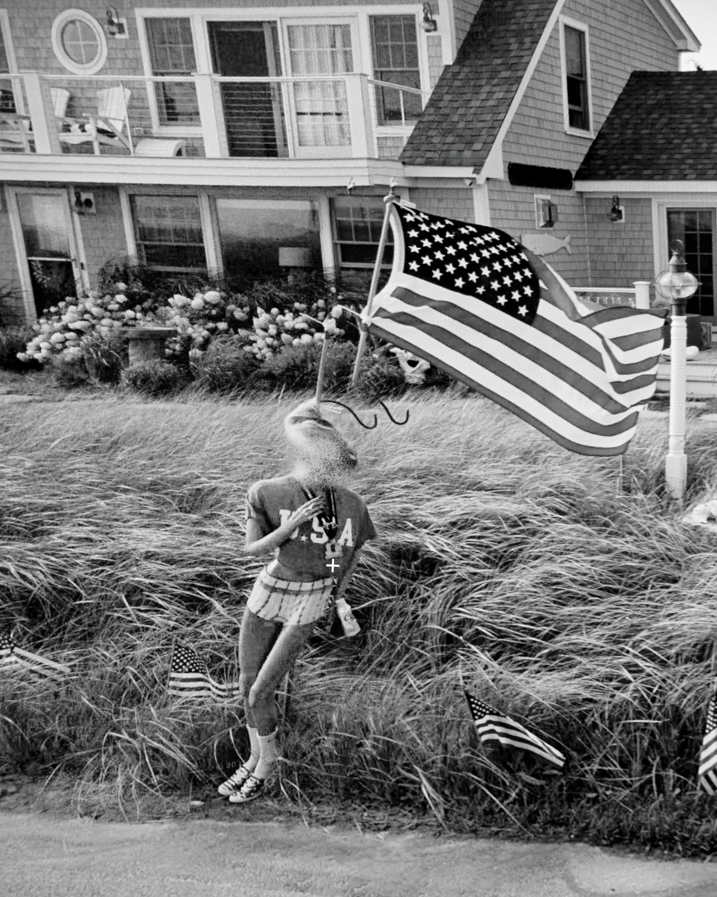 A child in a plaid skirt, striped socks, and a USA t-shirt running through tall grass in front of a house, with a large American flag-style beach umbrella nearby. The house has a balcony, multiple windows, and shingle siding.