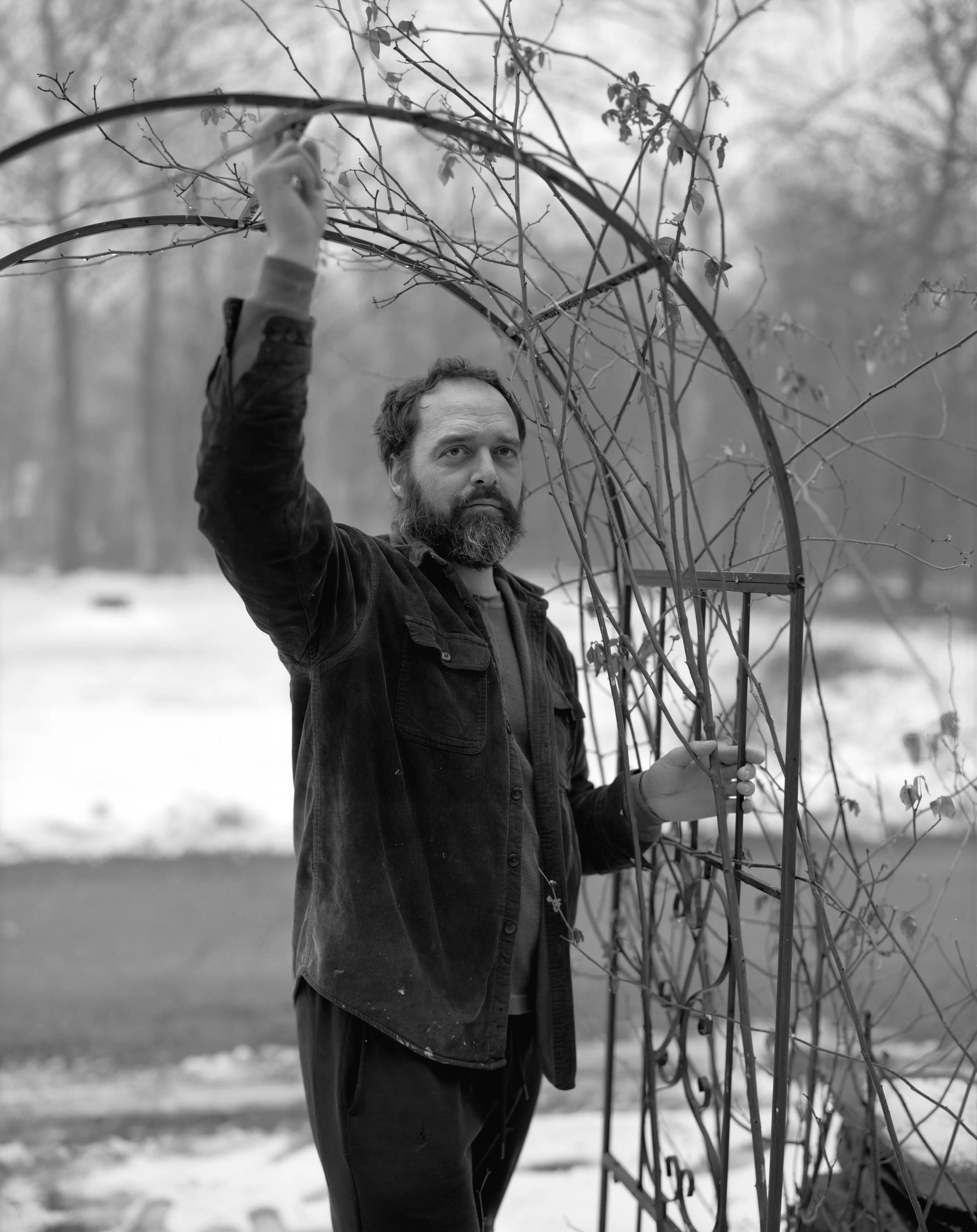 A man with a beard and curly hair holding a metal frame with entwined branches outdoors in a winter landscape