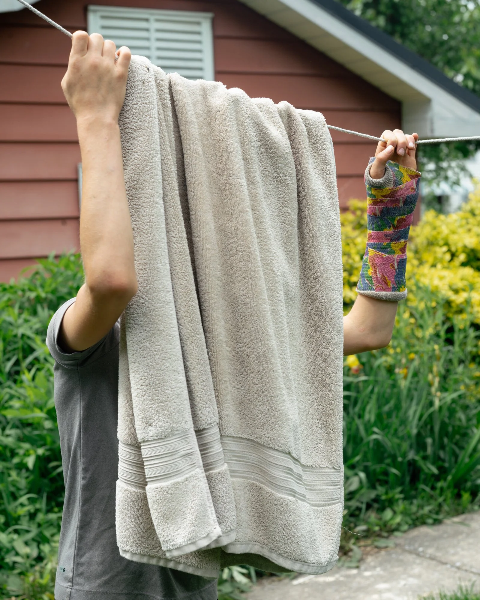 A person hanging a towel on a clothesline outside a house with red walls and white window shutters. The person's arm is visible, and they are wearing a gray shirt and a colorful, patterned sleeve on their other arm. Green plants and trees are in the 