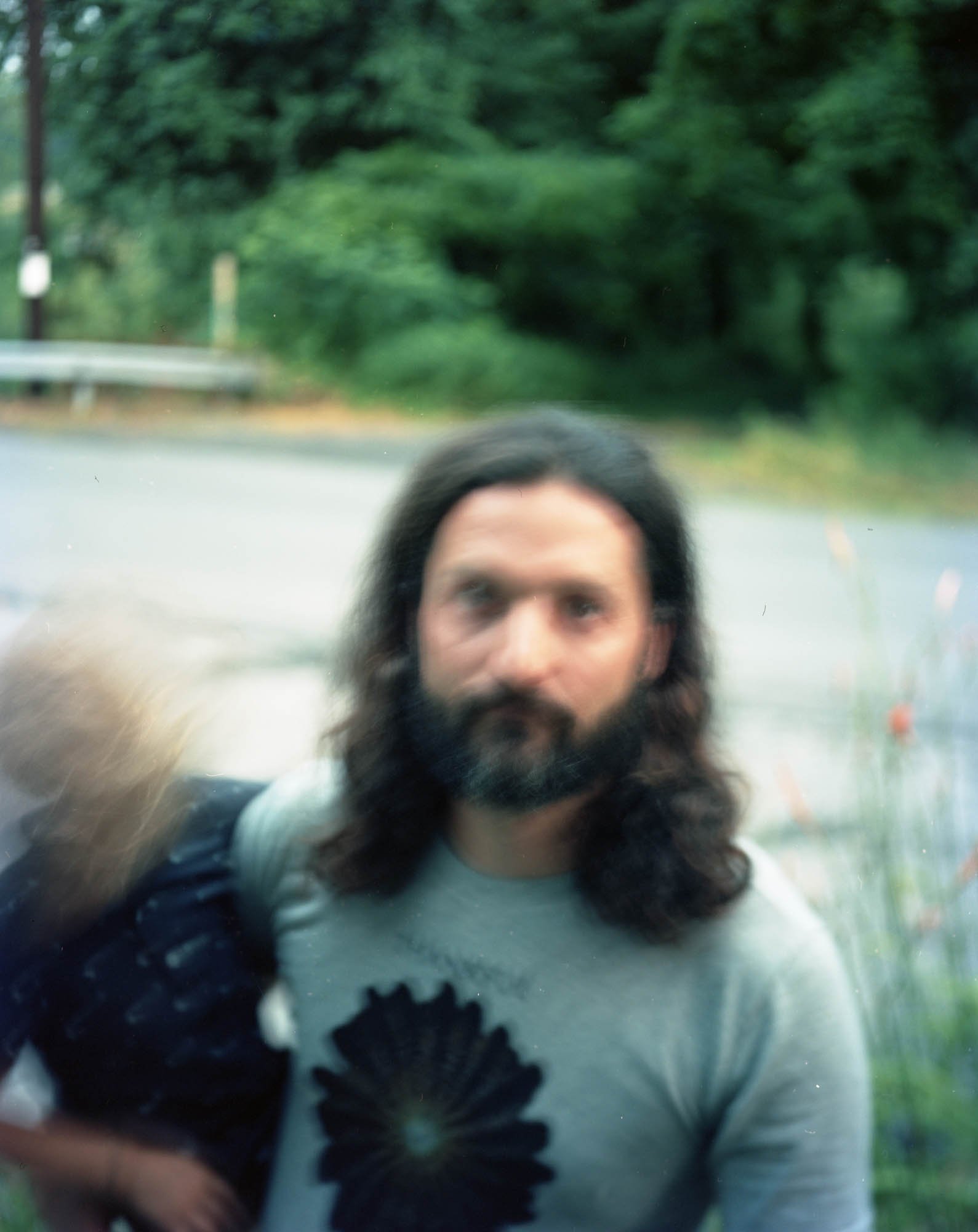 Self-portrait of a man with long dark hair and a beard, taking a photo through a reflective surface, with green trees and a road in the background.