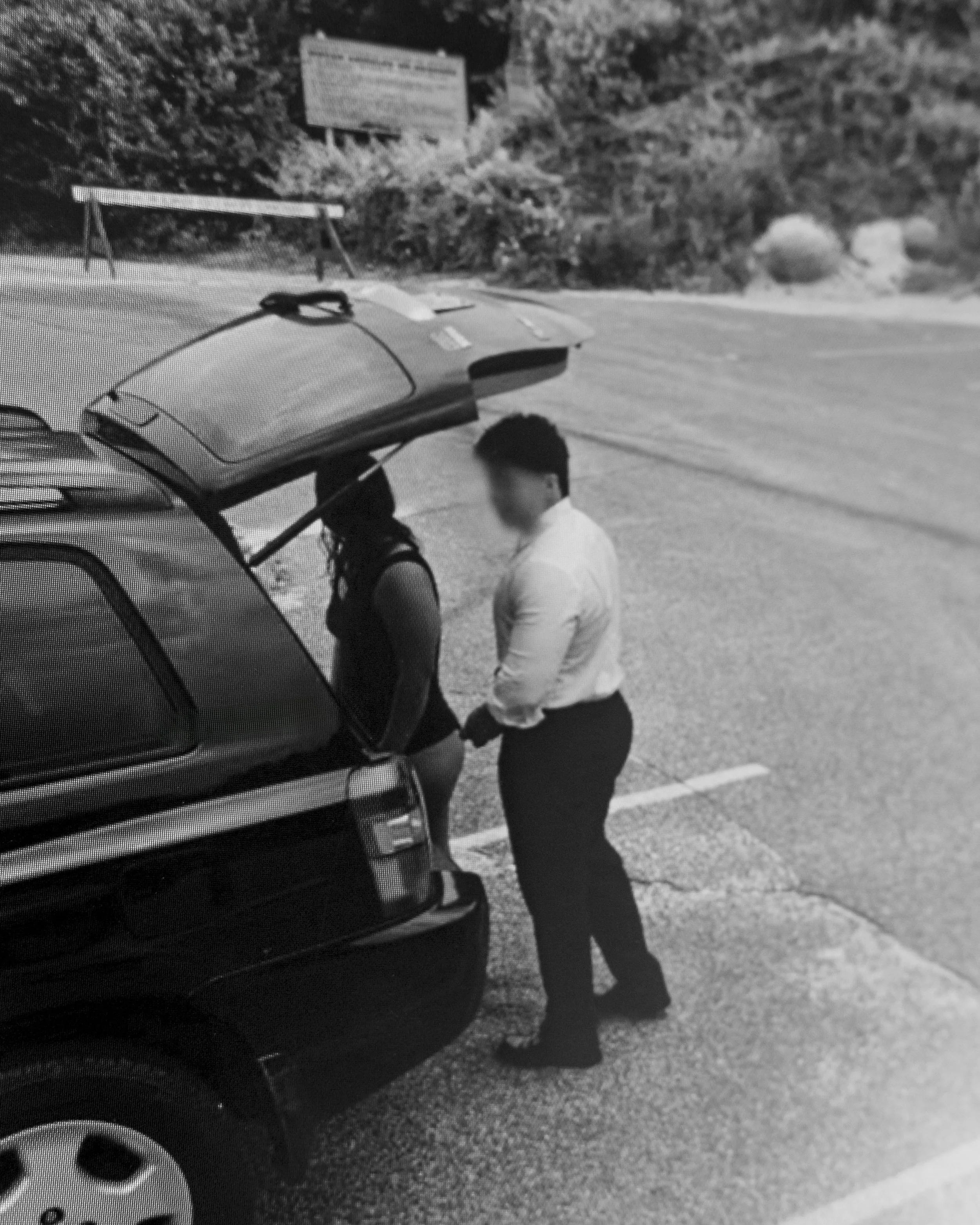 A black and white image of two people standing next to a car, with one person holding the car's door handle. The background includes a sign and some bushes or trees.