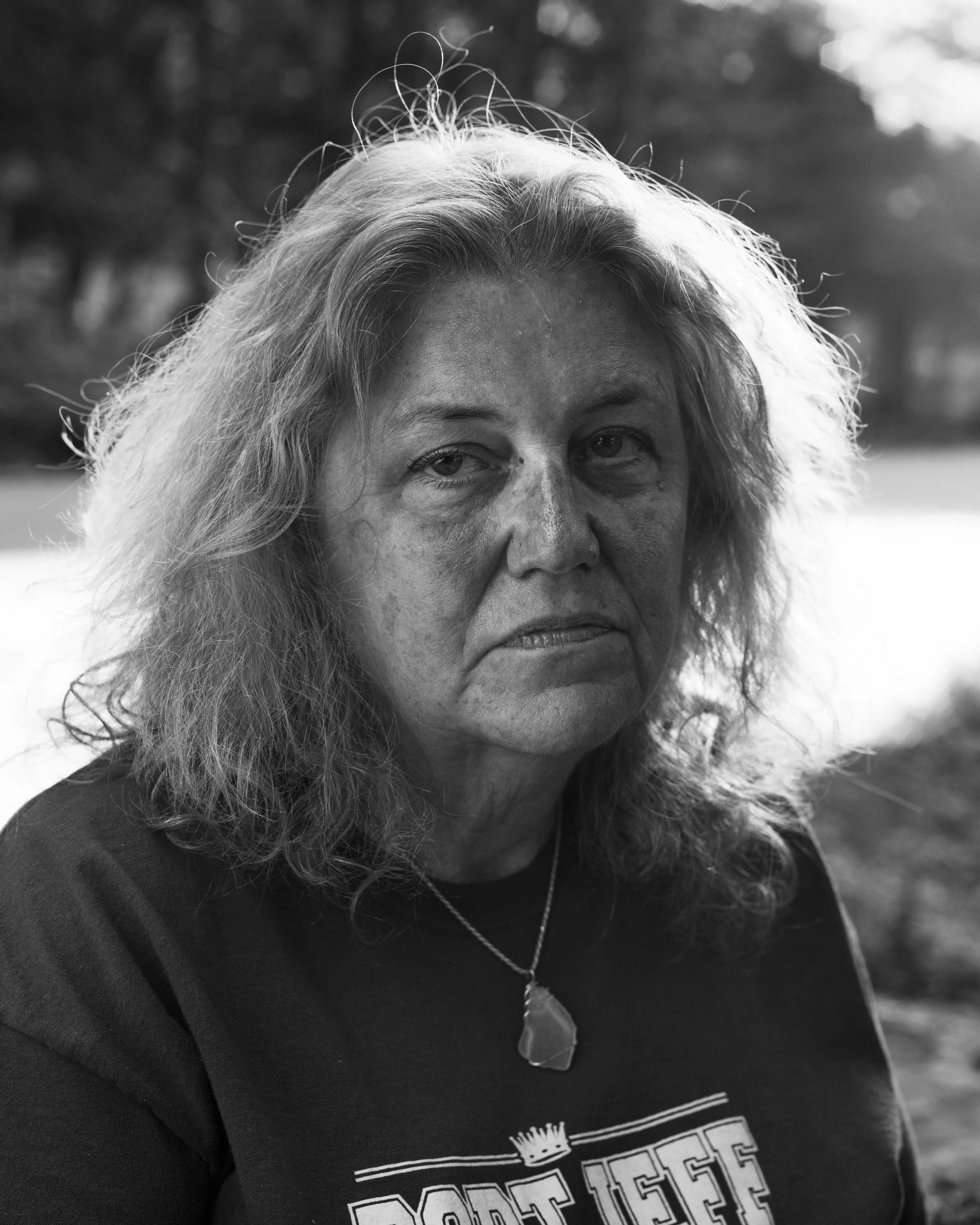 A woman with long, wavy gray hair, wearing a black shirt with white graphics, and a necklace with a leaf-shaped pendant.