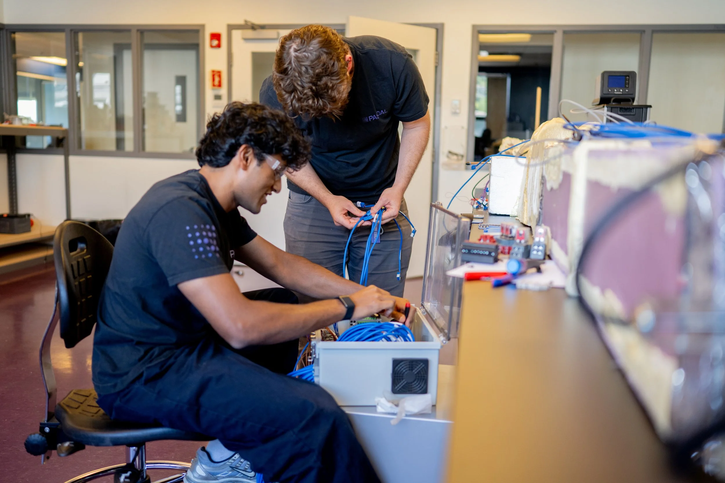 Two men working on electronic equipment at a workbench in a laboratory or classroom. One man is seated and working with tools, while the other is standing and connecting wires. The setting includes equipment, wires, and tools related to electronics or engineering.