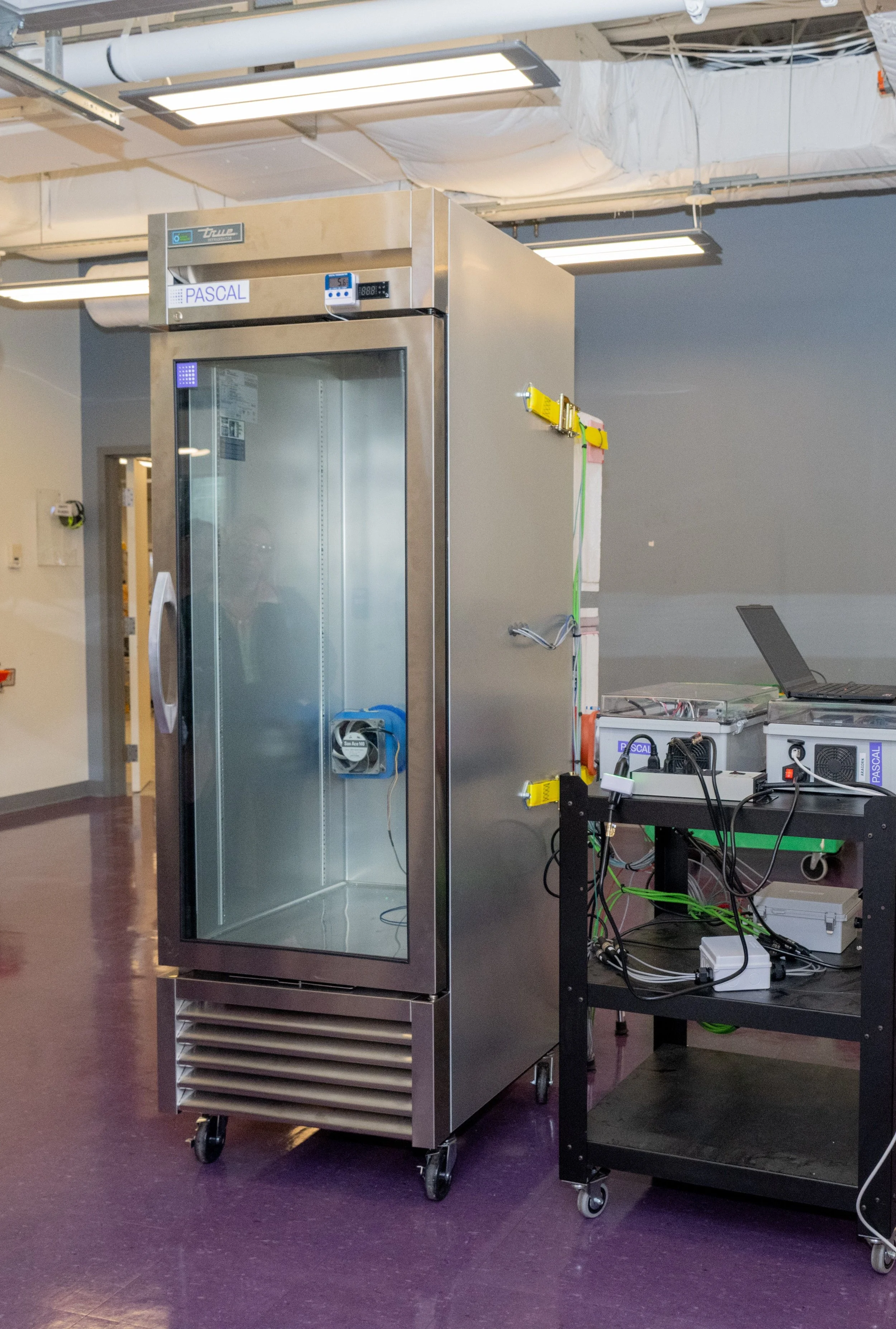 A stainless steel scientific or medical incubator with a glass door, placed on wheels in a laboratory or clinical setting. There are various electronic devices and wires on a black work table nearby, indicating ongoing testing or setup.