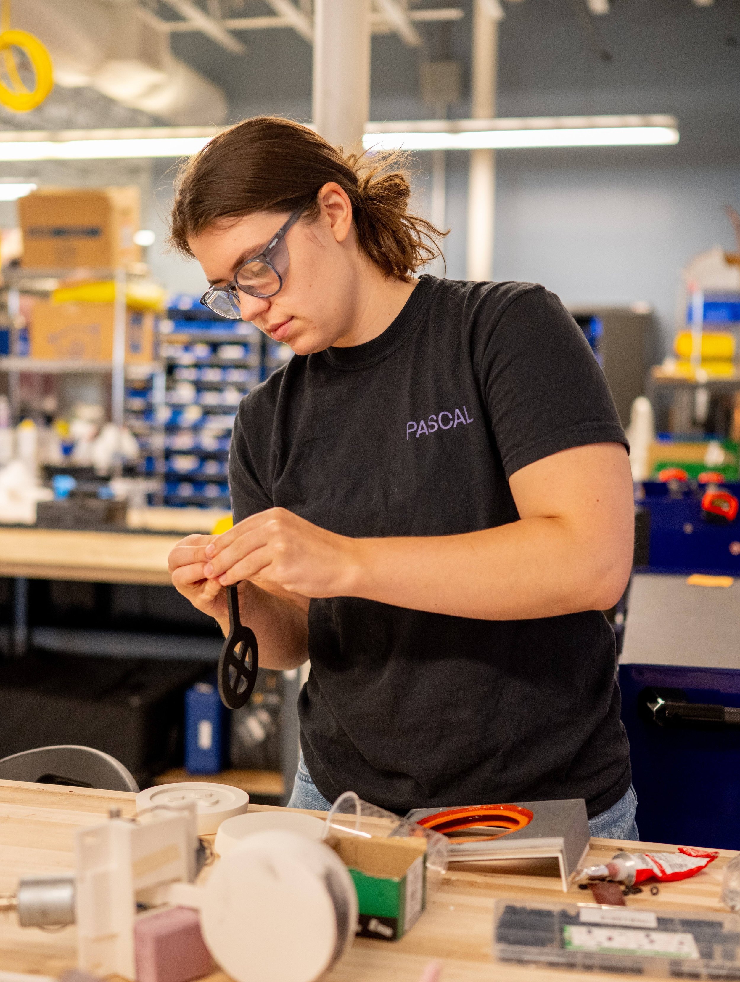 A woman with brown hair tied back, wearing glasses and a black T-shirt with the name 'PASCAL' on it, working at a cluttered wooden workbench in a workshop. She is holding a small object and appears focused. In the background, there are shelves with various boxes and tools, illuminated by fluorescent lighting.