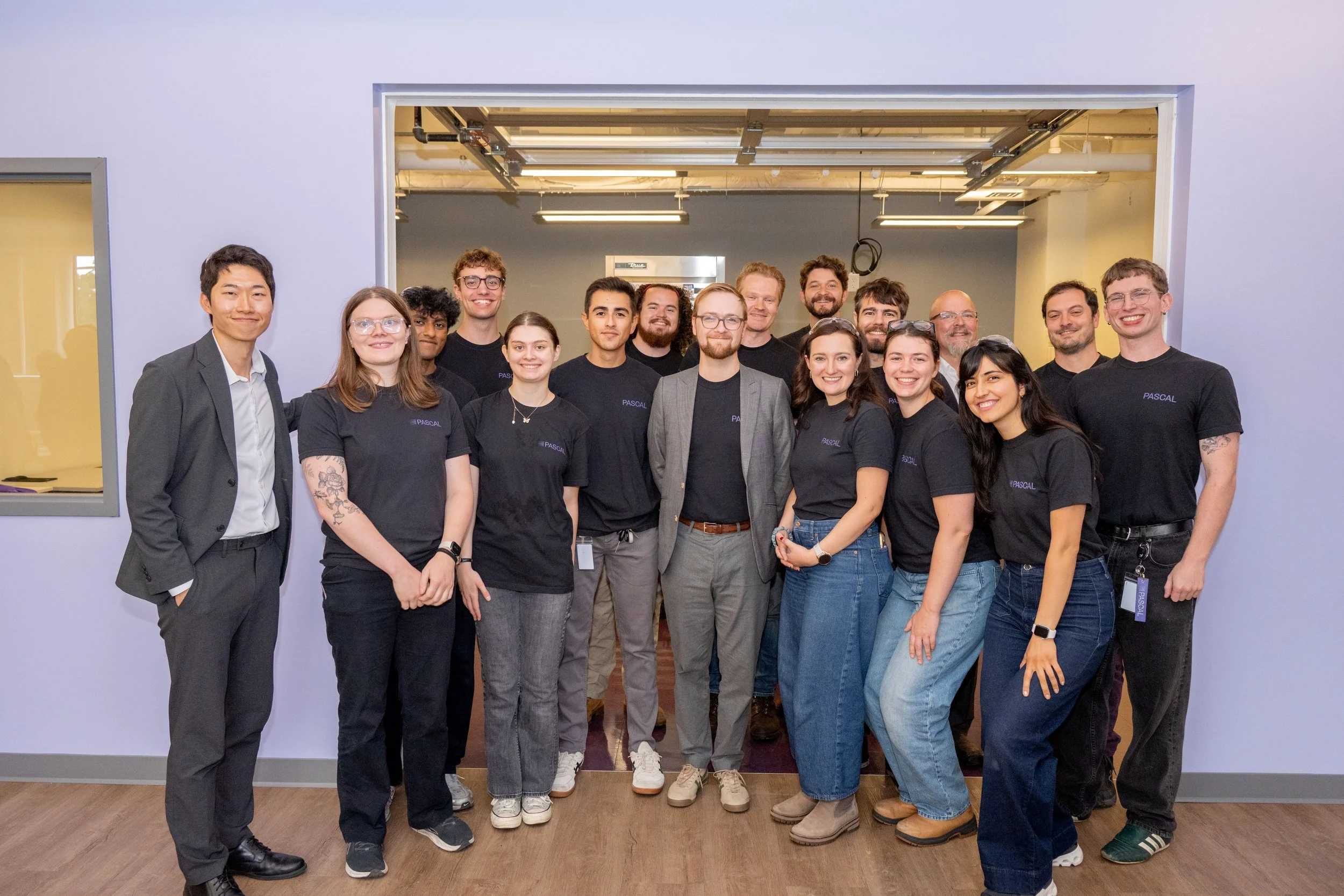 Group of diverse young adults and older professionals standing together in an office or industrial space, smiling for a photo.