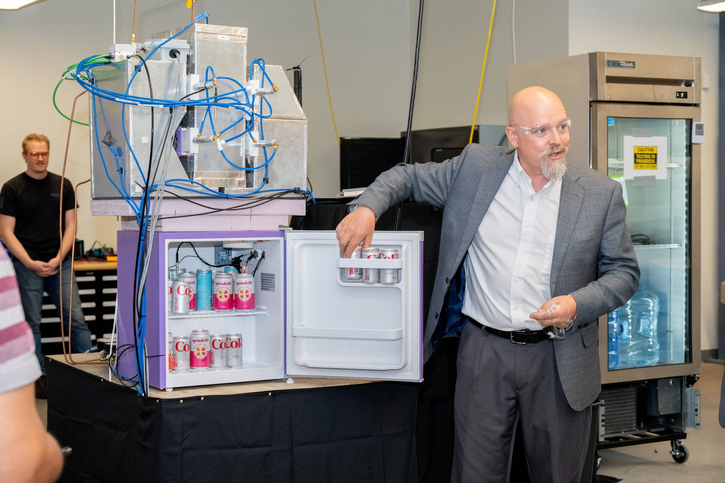 A man in a gray suit and glasses standing next to a purple mini fridge filled with cans of CoCola, at an event with people and equipment in the background.