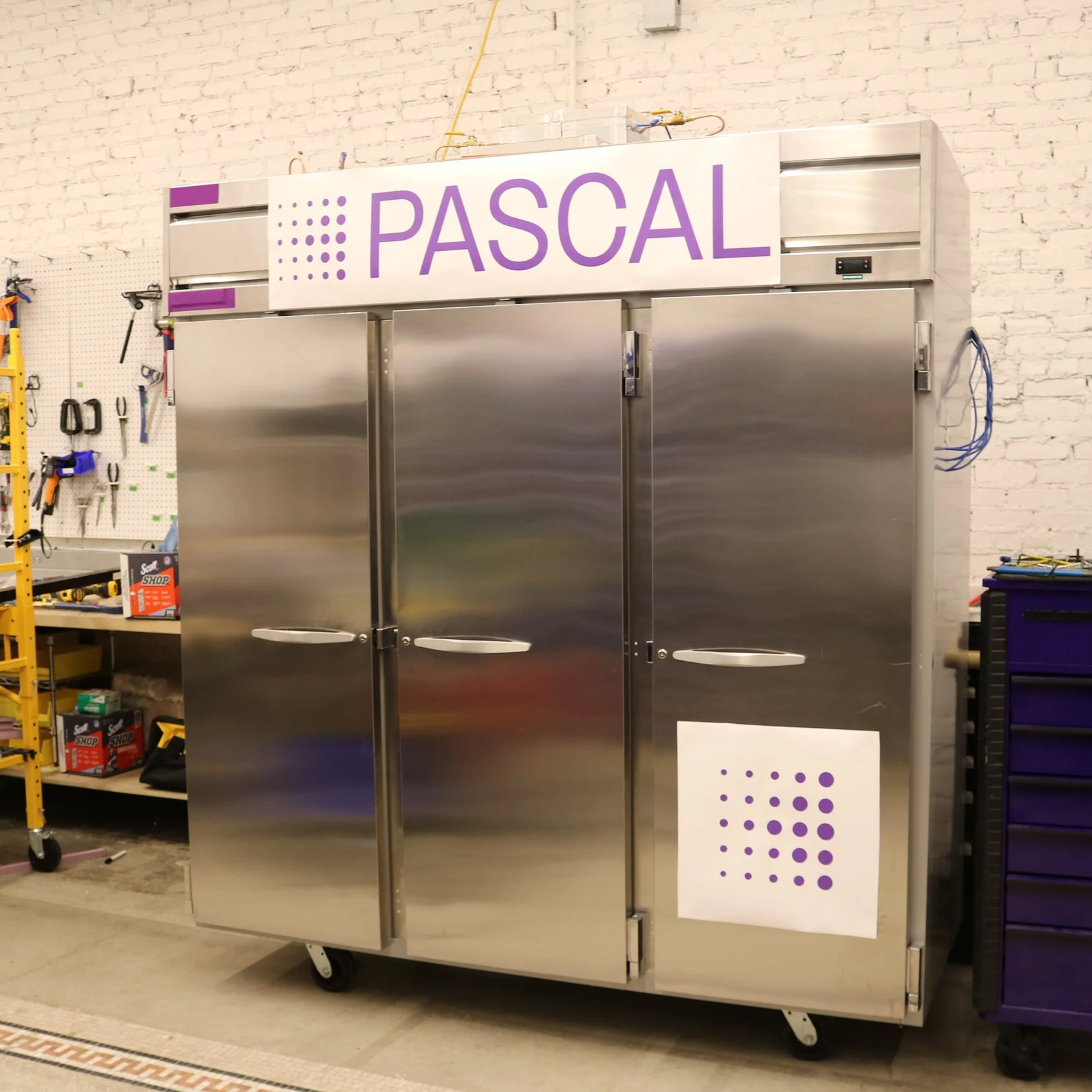 A large stainless steel refrigerator with a sign on top reading 'PASCAL' in purple capital letters. The refrigerator is in a workshop with tools hanging on a pegboard on the left and other equipment around.