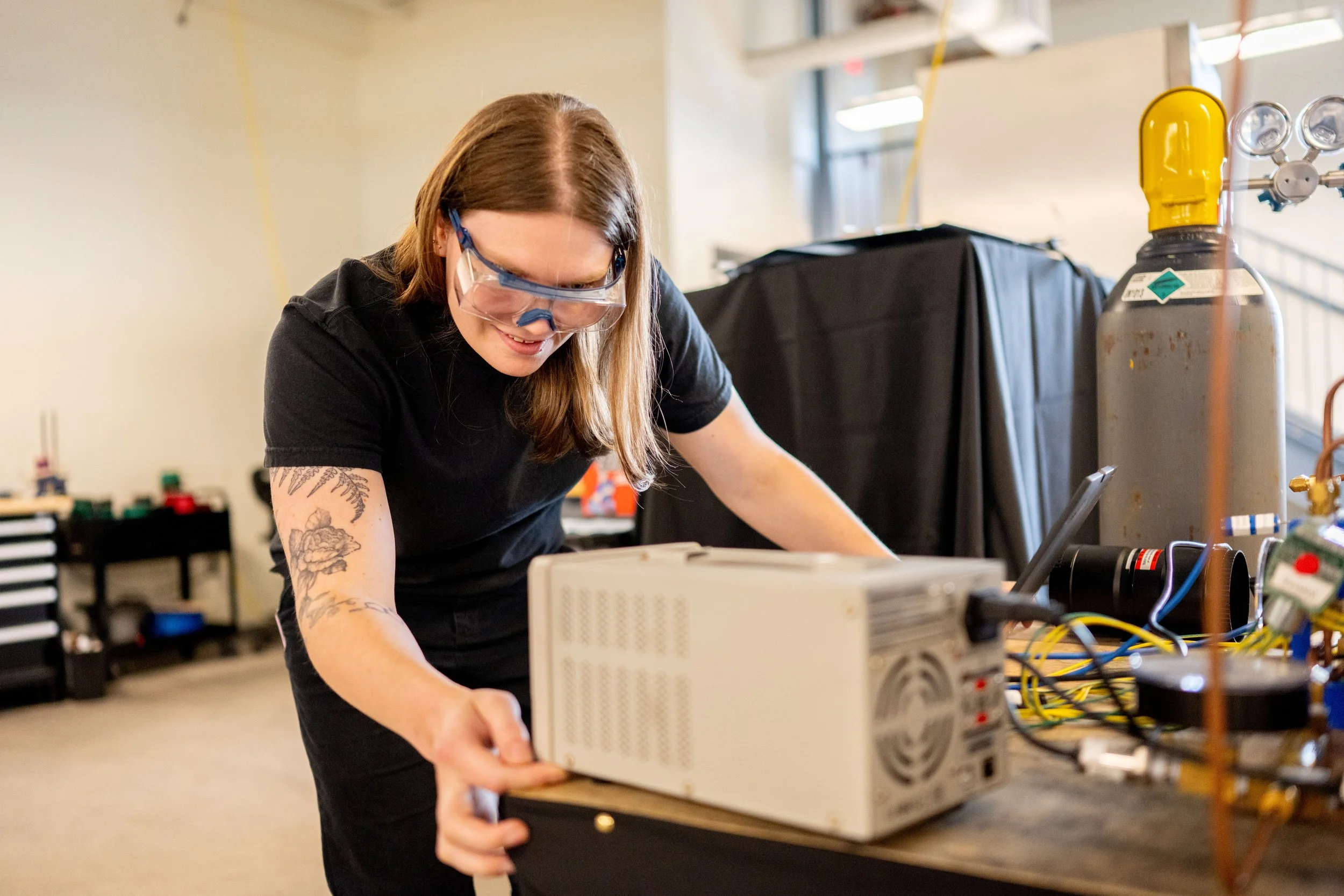 A woman with long hair, glasses, and tattoos on her arm is working with electronic equipment and wires on a worktable in an industrial or workshop setting.