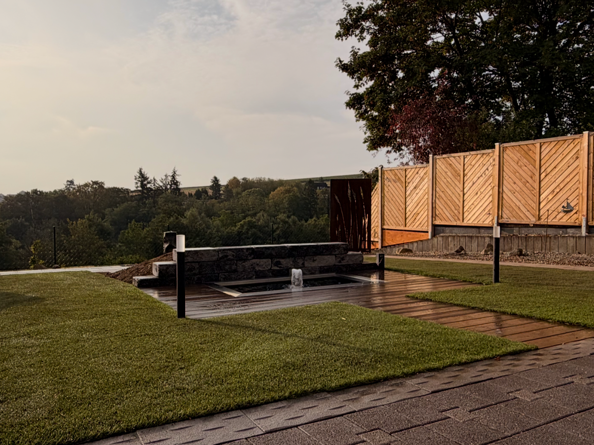 Garten mit Wasserfall, Holzplanken, Rasenflächen und Holzzaun im Hintergrund, umgeben von Bäumen