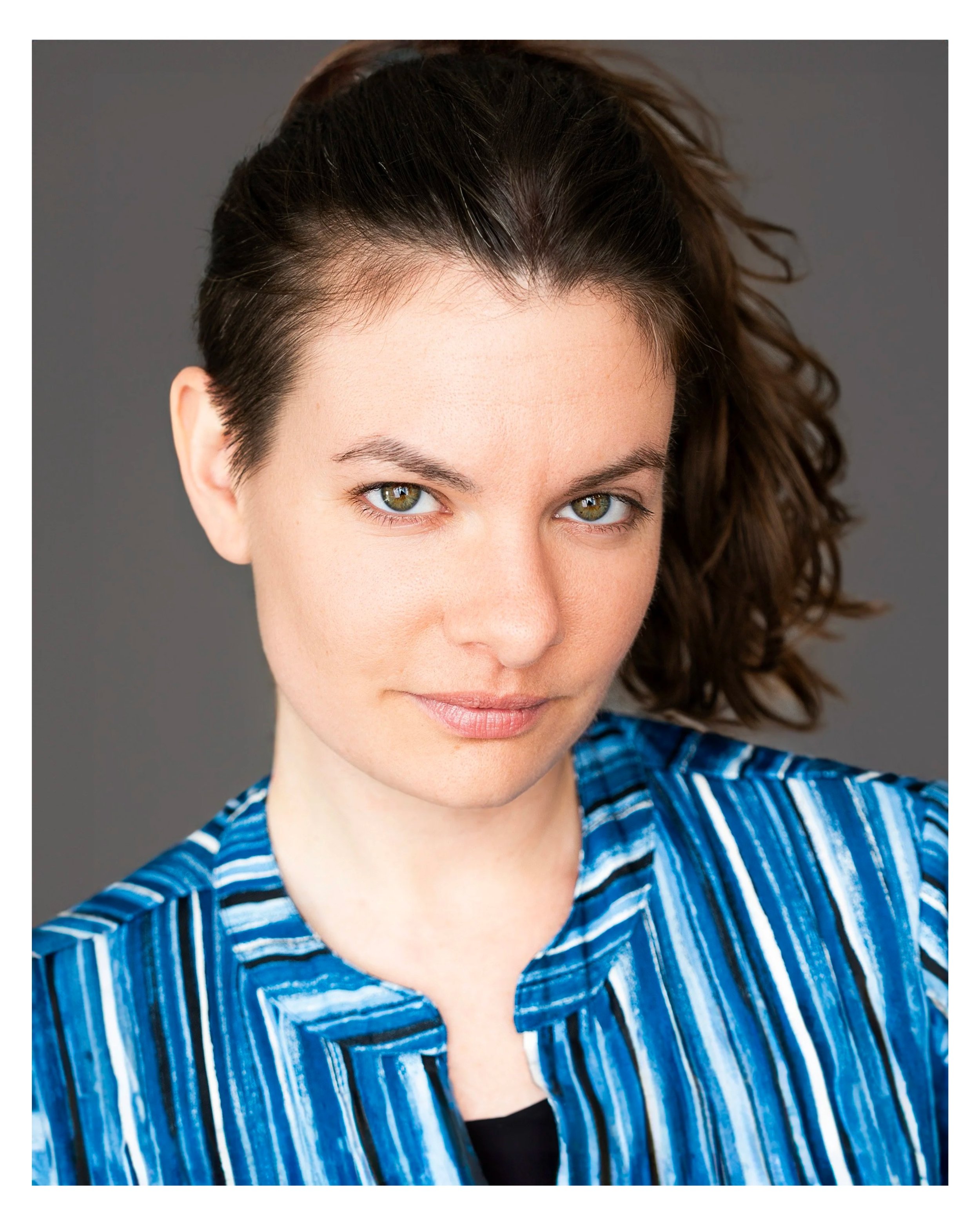 Close-up of a woman with dark hair, hazel eyes, and fair skin, wearing a blue and white striped shirt, against a grey background.