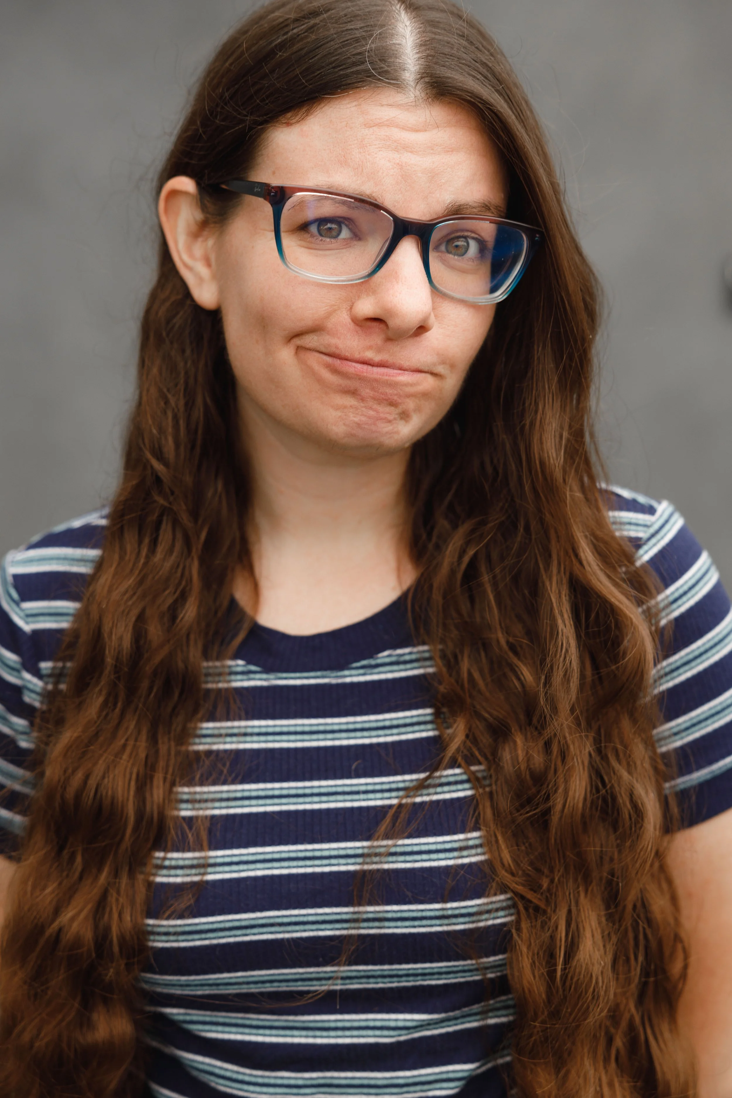Close-up of a woman with long wavy brown hair, wearing glasses and a navy blue and white striped shirt, displaying a subtle smirk against a plain gray background.