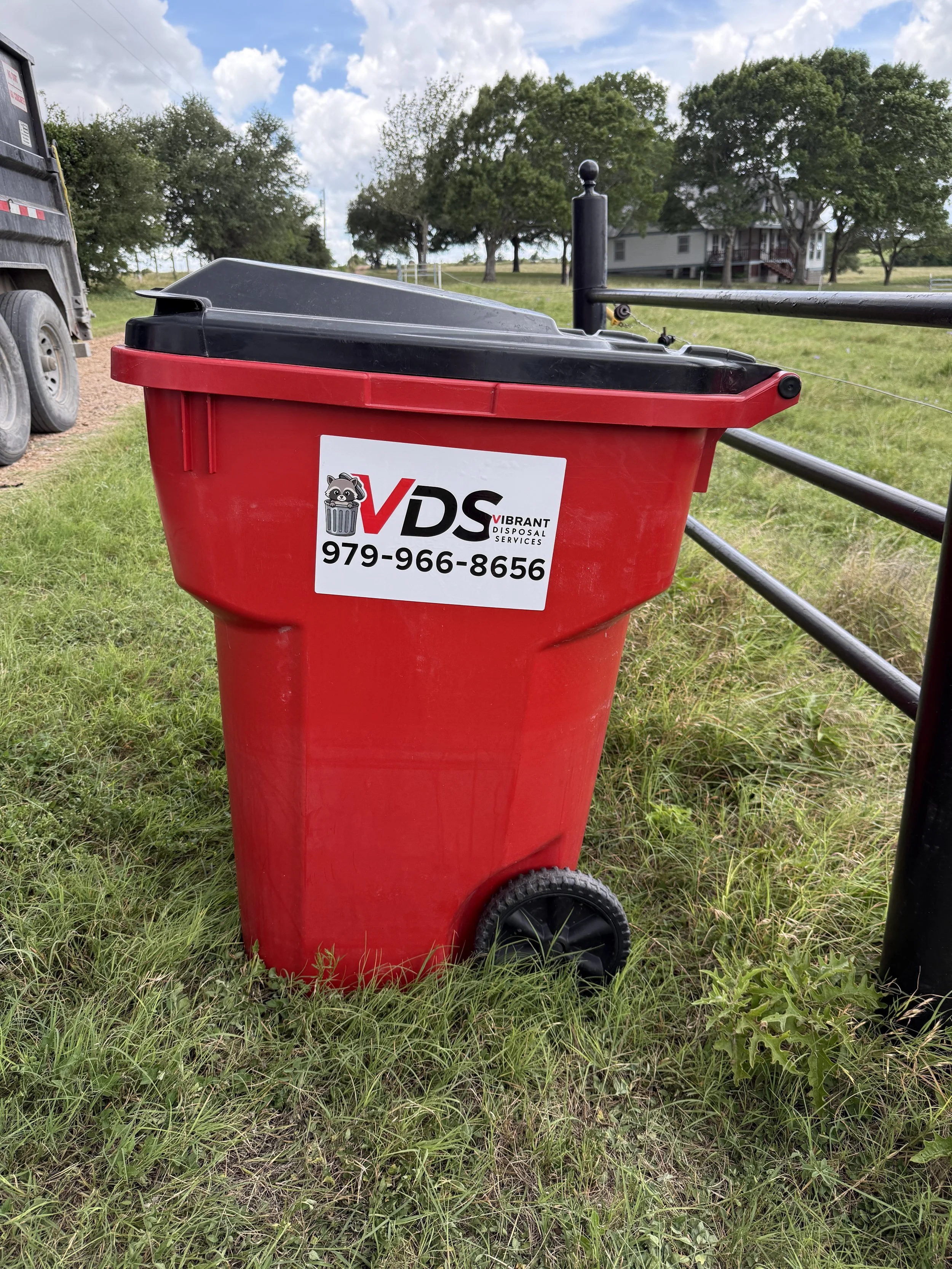 Red trash bin with a black lid and a sticker with a raccoon logo, the text 'VDS Vibrant Disposal Services', and a phone number, standing on grass with a black gate, trees, a house, and a cloudy sky in the background.