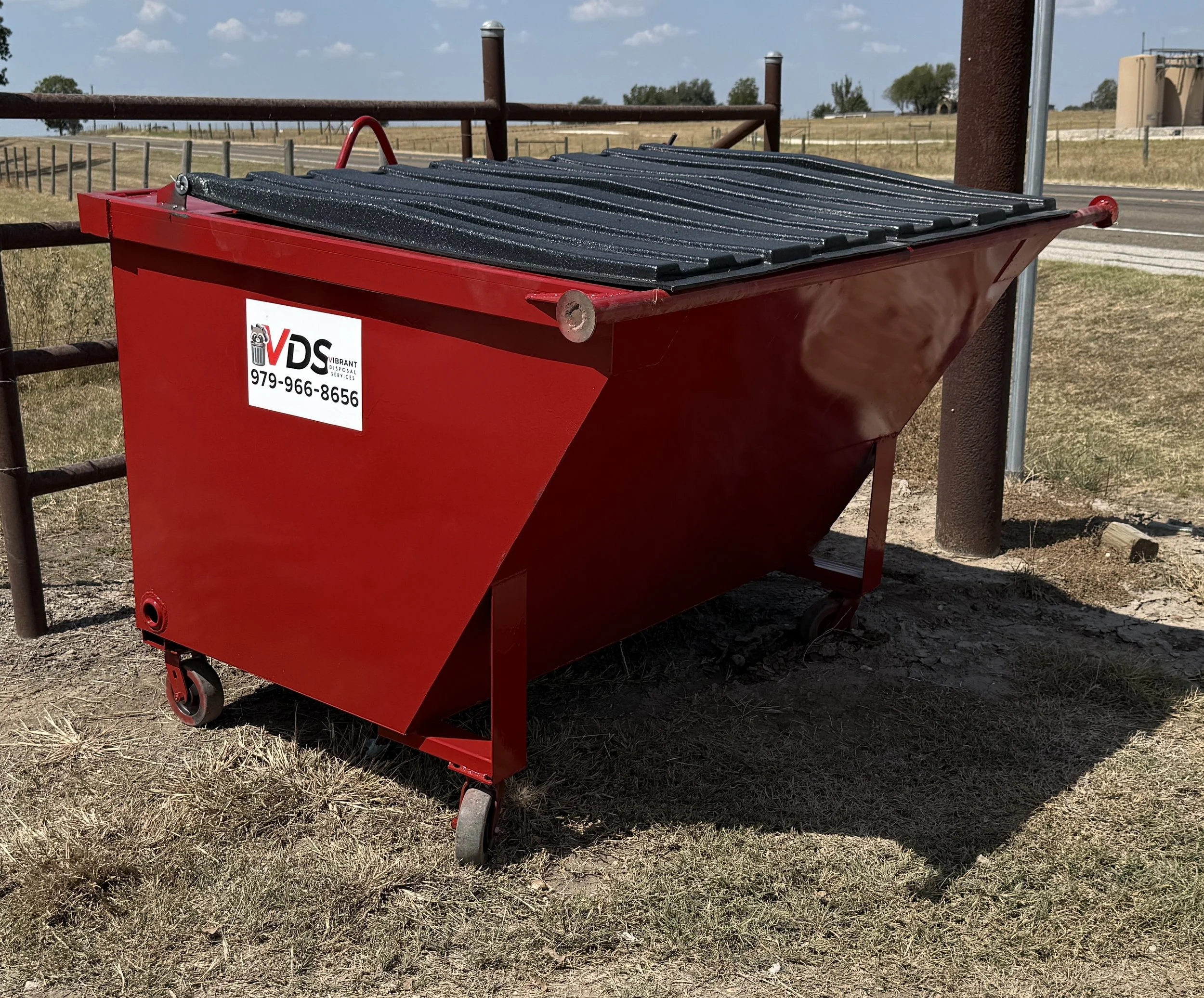 A red dumpster with black lid, situated outdoors on a patch of dry grass, with a rural landscape in the background. The dumpster has a white label with the logo 'VDS' and a phone number.