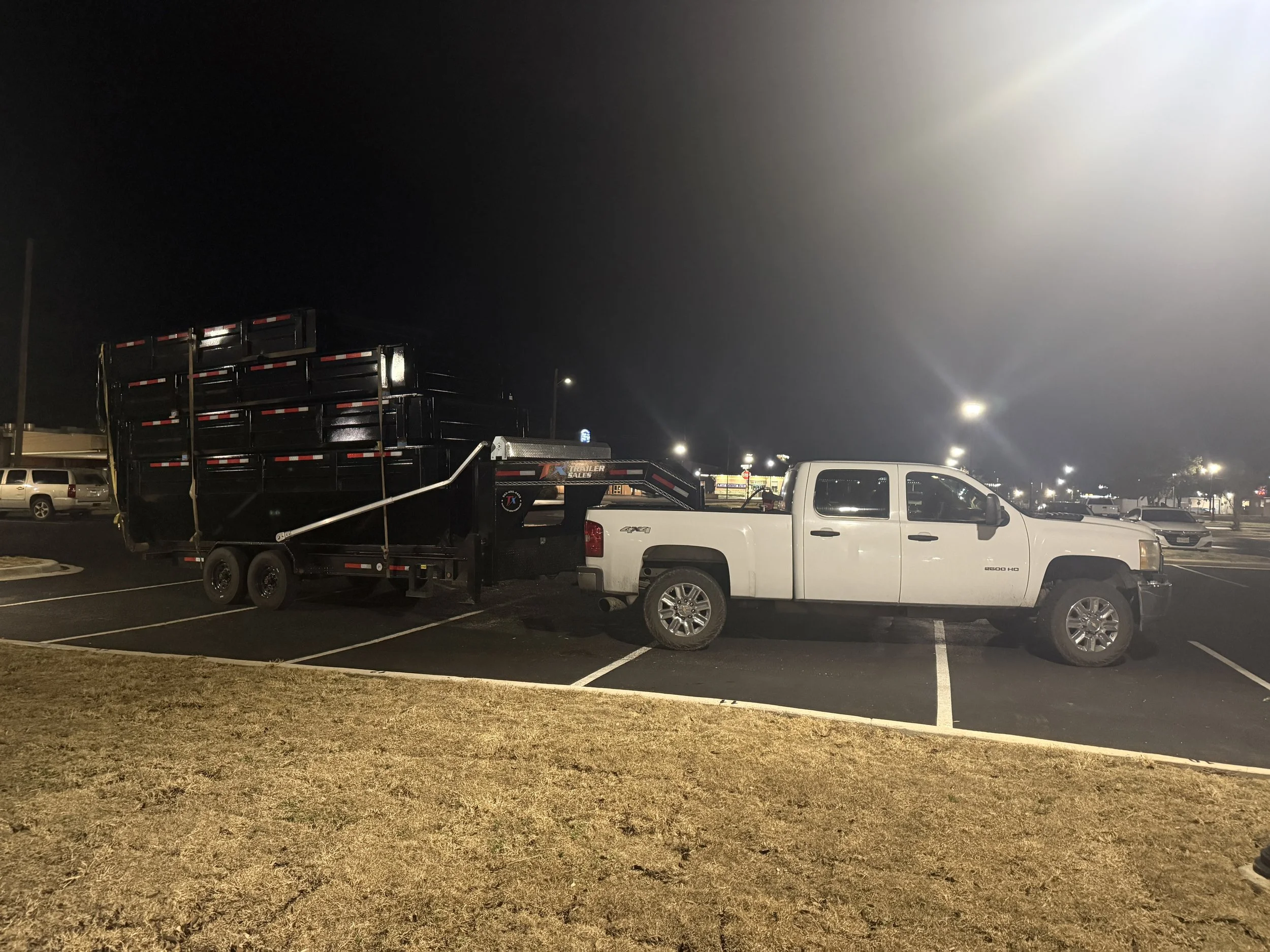 A parking lot at night with several vehicles including a white pickup truck towing a flatbed trailer, and other cars parked in the background. There are streetlights illuminating the scene and a dark sky above.