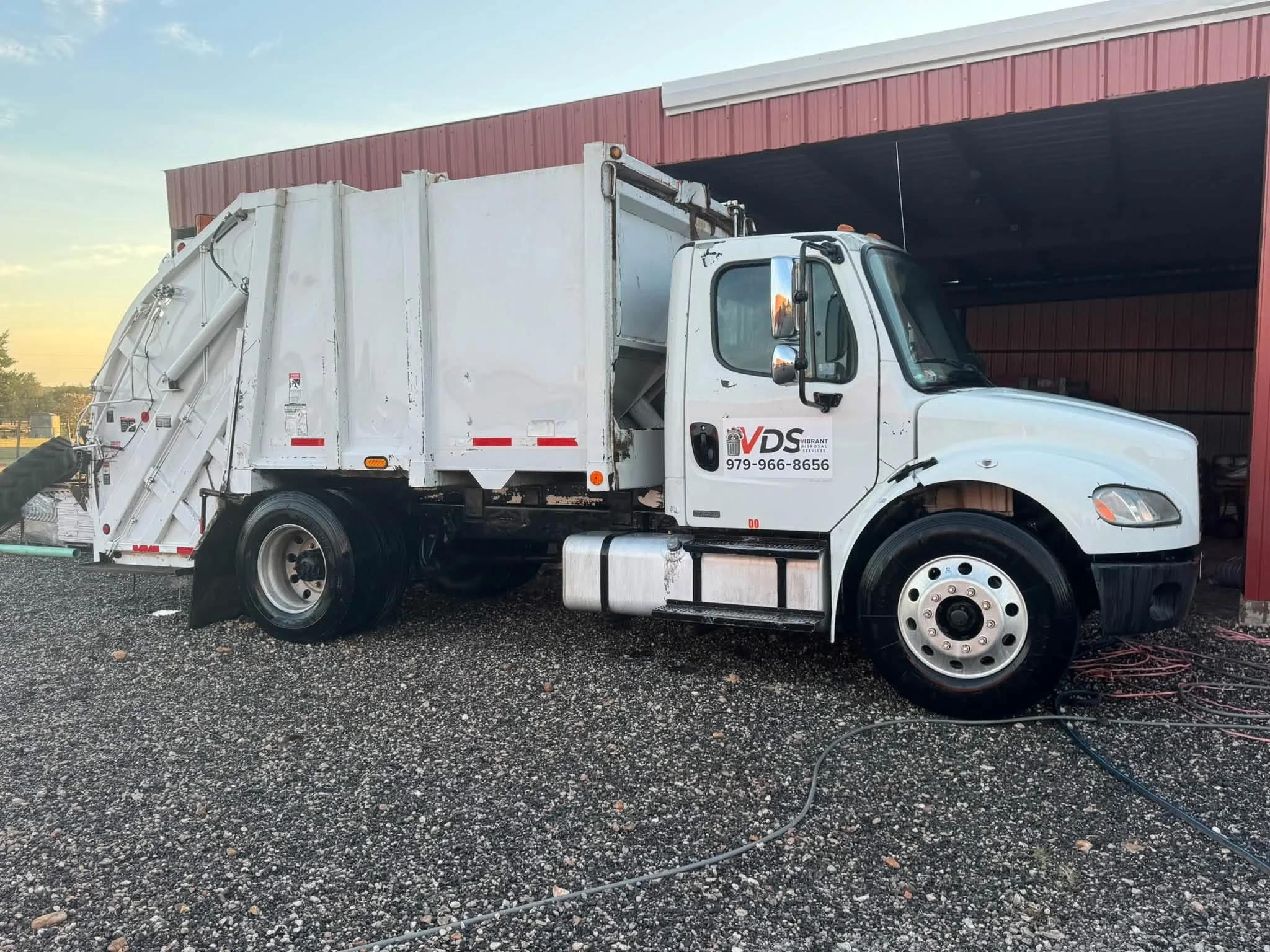 A white garbage truck parked outside a red metal building on gravel ground with some electrical wires in the foreground.