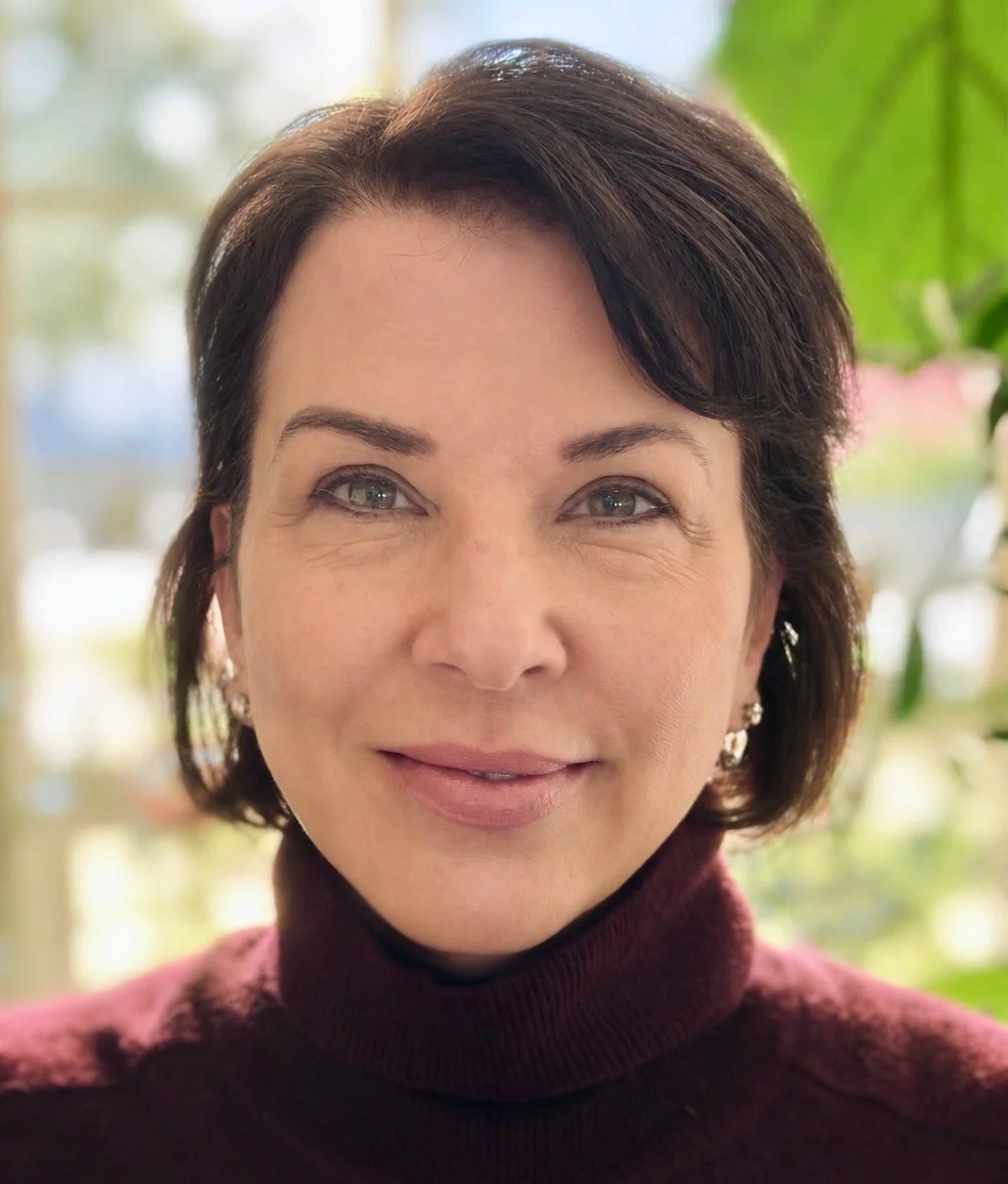 A woman with short brown hair and hazel green eyes, wearing a maroon turtleneck sweater and stud earrings, standing in front of a background with green leaves.