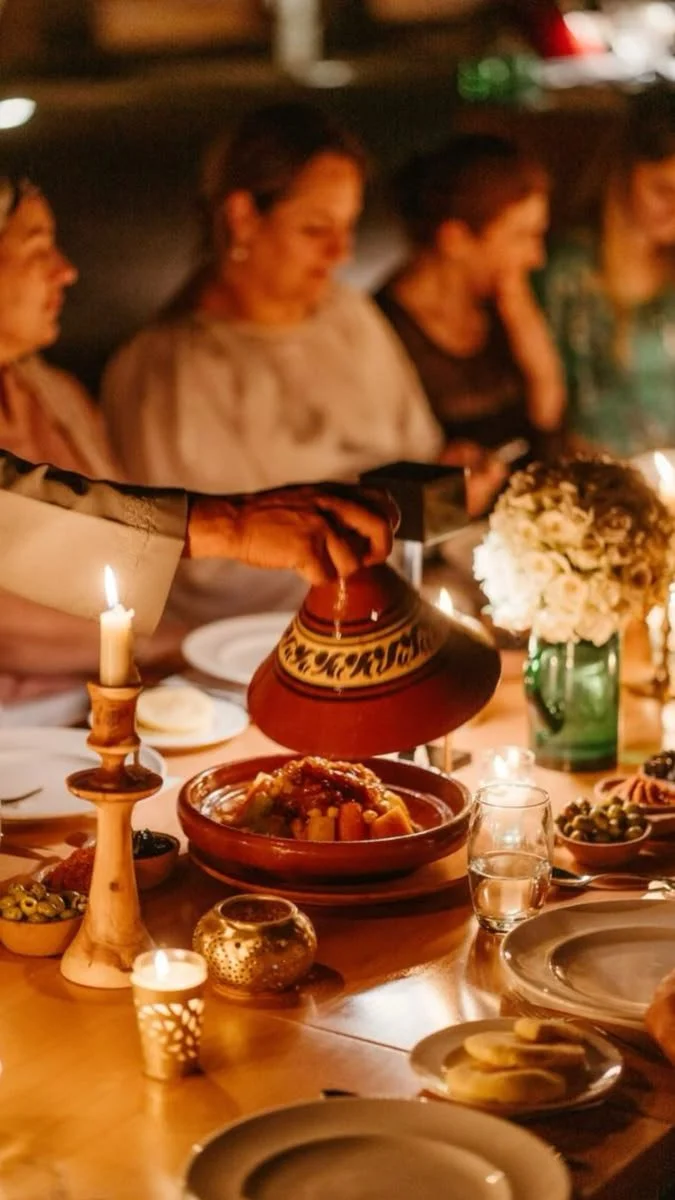 Friends gathered around a candlelit dinner table with Moroccan tagine, shared dishes, wine glasses, and a flower centerpiece