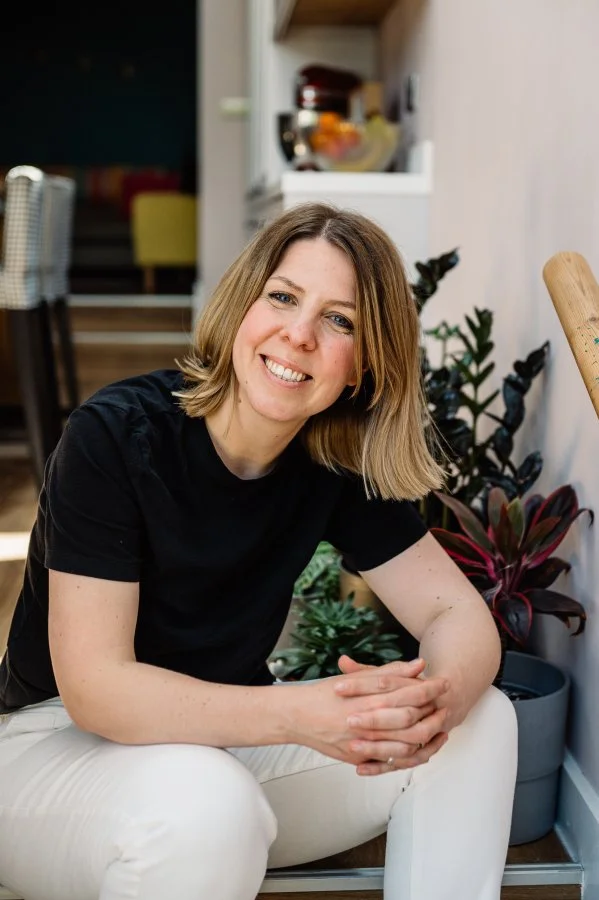 A woman with shoulder-length blonde hair smiling at the camera, sitting in a cozy indoor space with plants and a kitchen in the background.