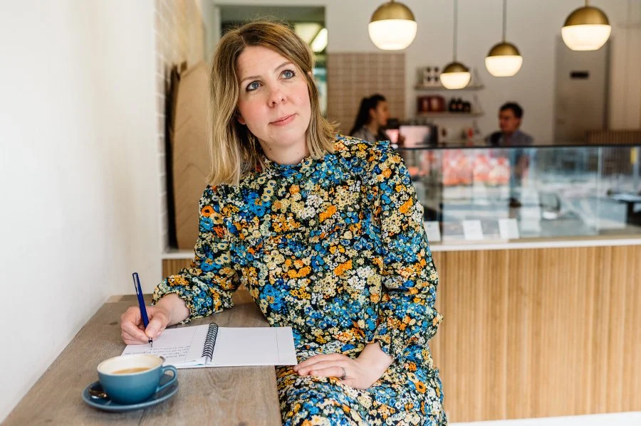 A woman with shoulder-length blond hair wearing a colorful floral dress sitting at a wooden table in a coffee shop, writing in a notebook with a blue pen, with a cup of coffee beside her. In the background, there are three people sitting at a table inside the shop.