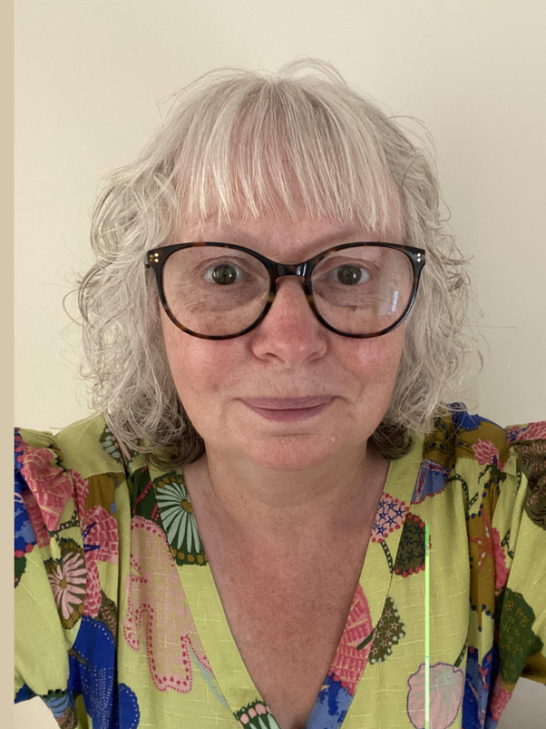 A woman with curly gray hair and black glasses looking at the camera, wearing a colorful floral shirt against a plain white wall background.