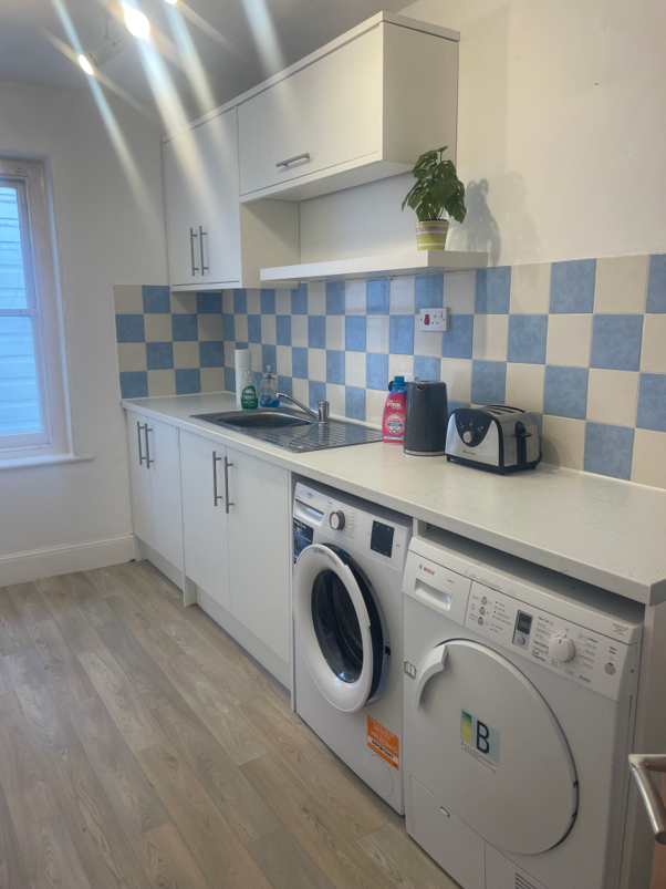 Kitchen with white cabinets and blue checkered tile backsplash. Contains a sink, dish soap, kettle, toaster, and a potted plant on a shelf. There is a washing machine and dryer under the countertop, a window on the left, and wood laminate flooring.