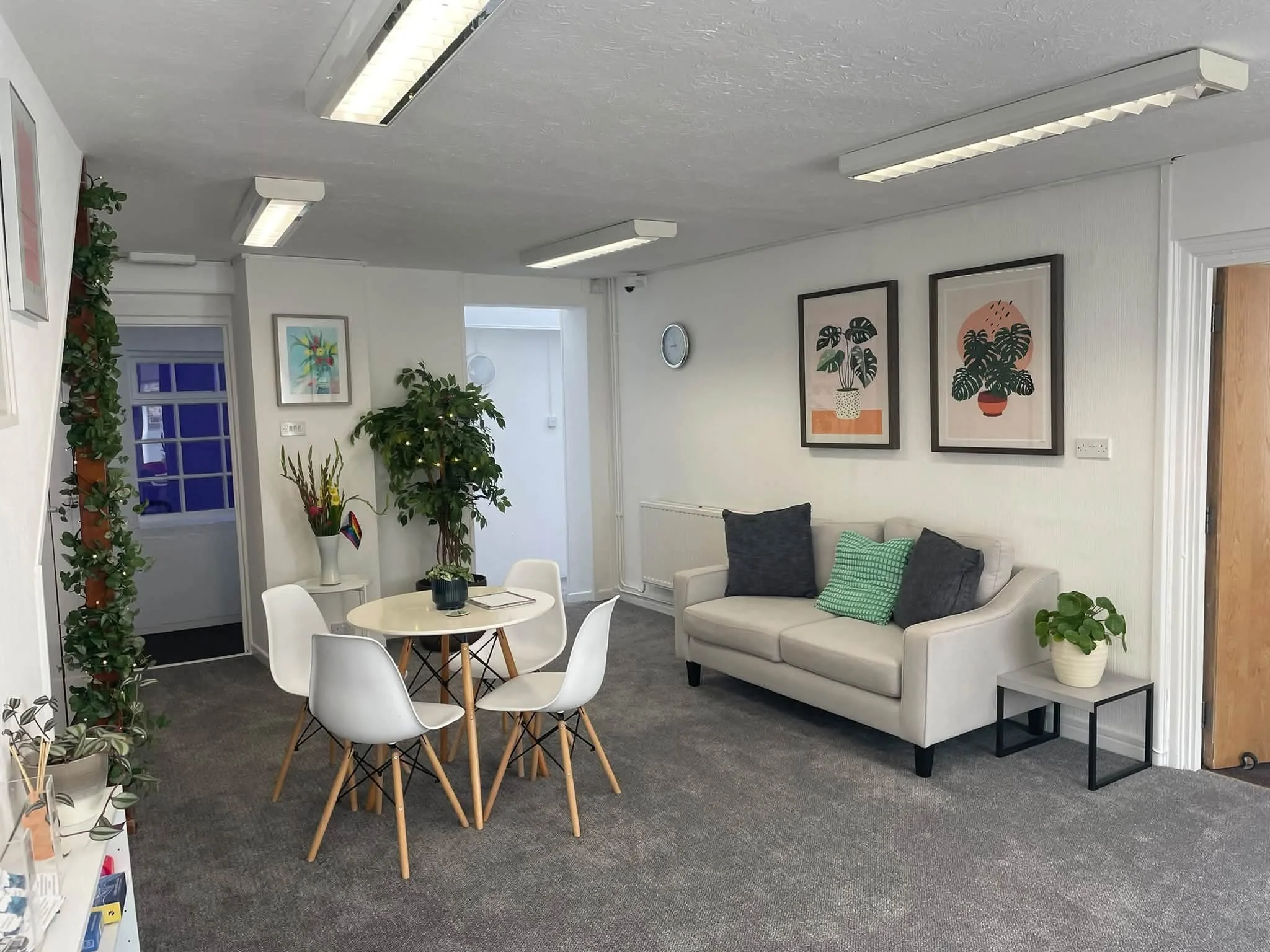 An indoor office or lounge area with white walls, gray carpet, and wooden accents. There is a round white table with white chairs, a white sofa with green and gray pillows, framed art on the walls featuring plants, several potted plants, and modern lighting fixtures on the ceiling.