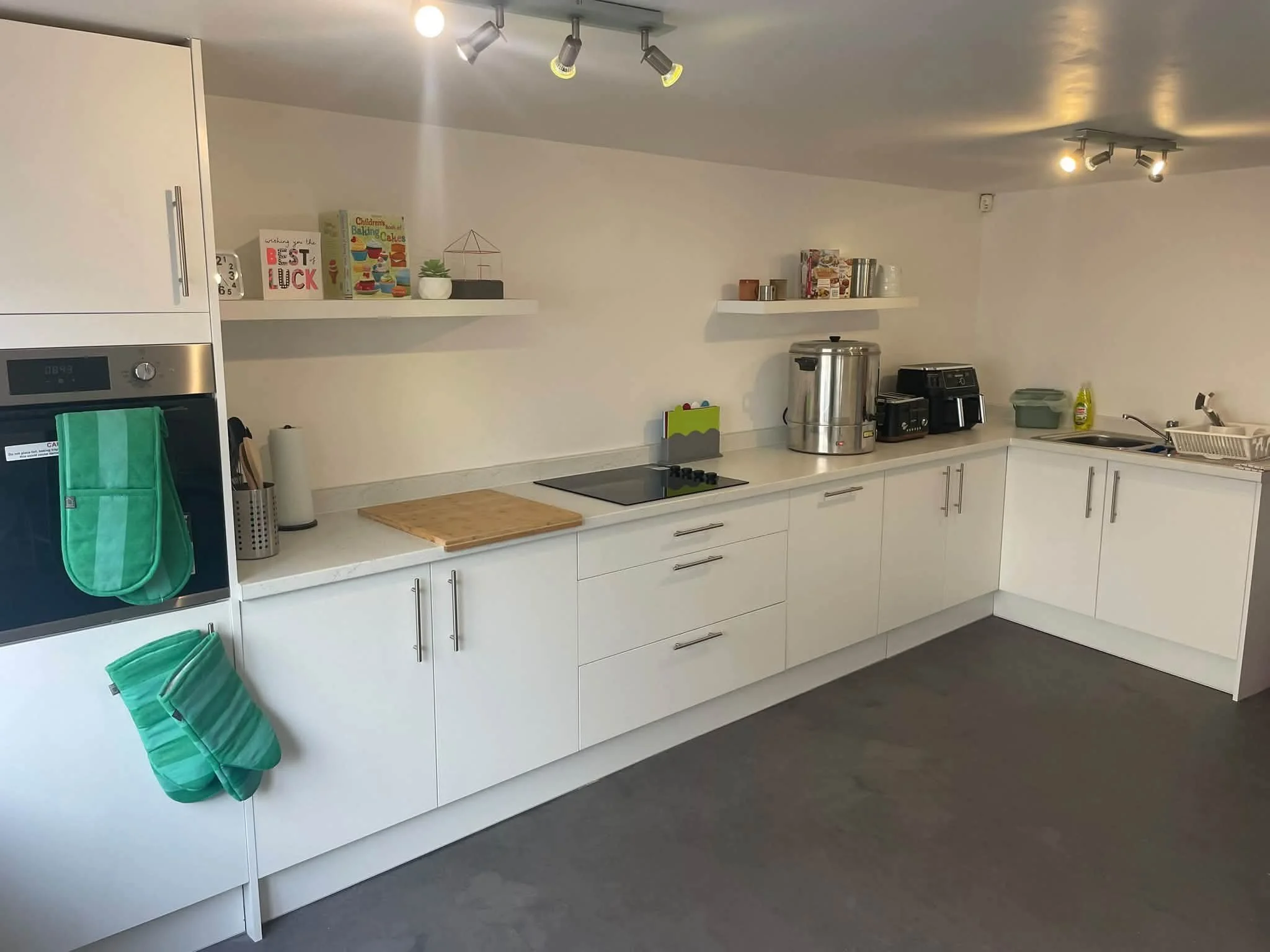 Modern kitchen with white cabinets, gray countertops, and black flooring. There are two shelves with decorative items, a stove with a cutting board, various small appliances, and cleaning supplies. Two green oven mitts are hanging on the side of the cabinets.