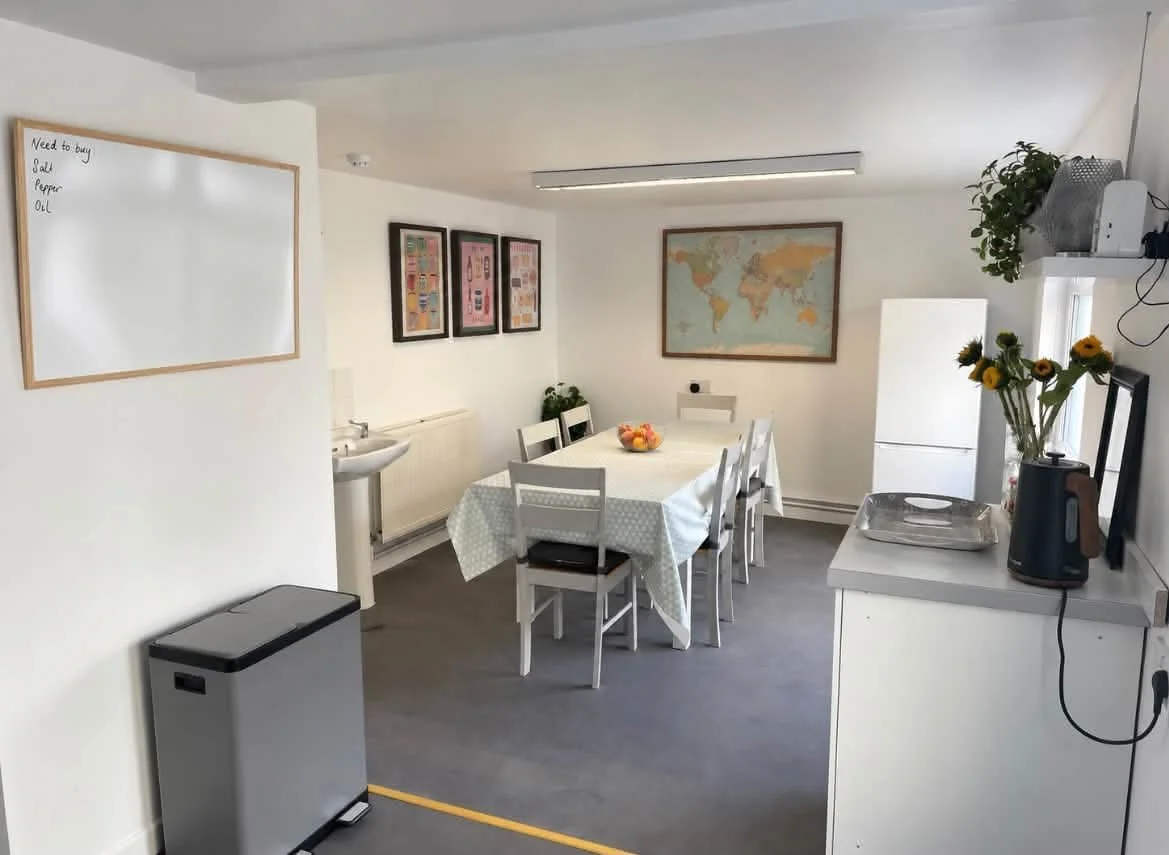 A dining area with a rectangular table covered with a white checkered tablecloth, surrounded by six white chairs, and a bowl of fruit in the center. There are framed posters on the wall, a world map, and a small sink with a potted plant nearby. To the right, a white refrigerator and a kitchen counter with a vase of yellow flowers, a kettle, and a metal tray. A whiteboard on the wall has notes written on it.