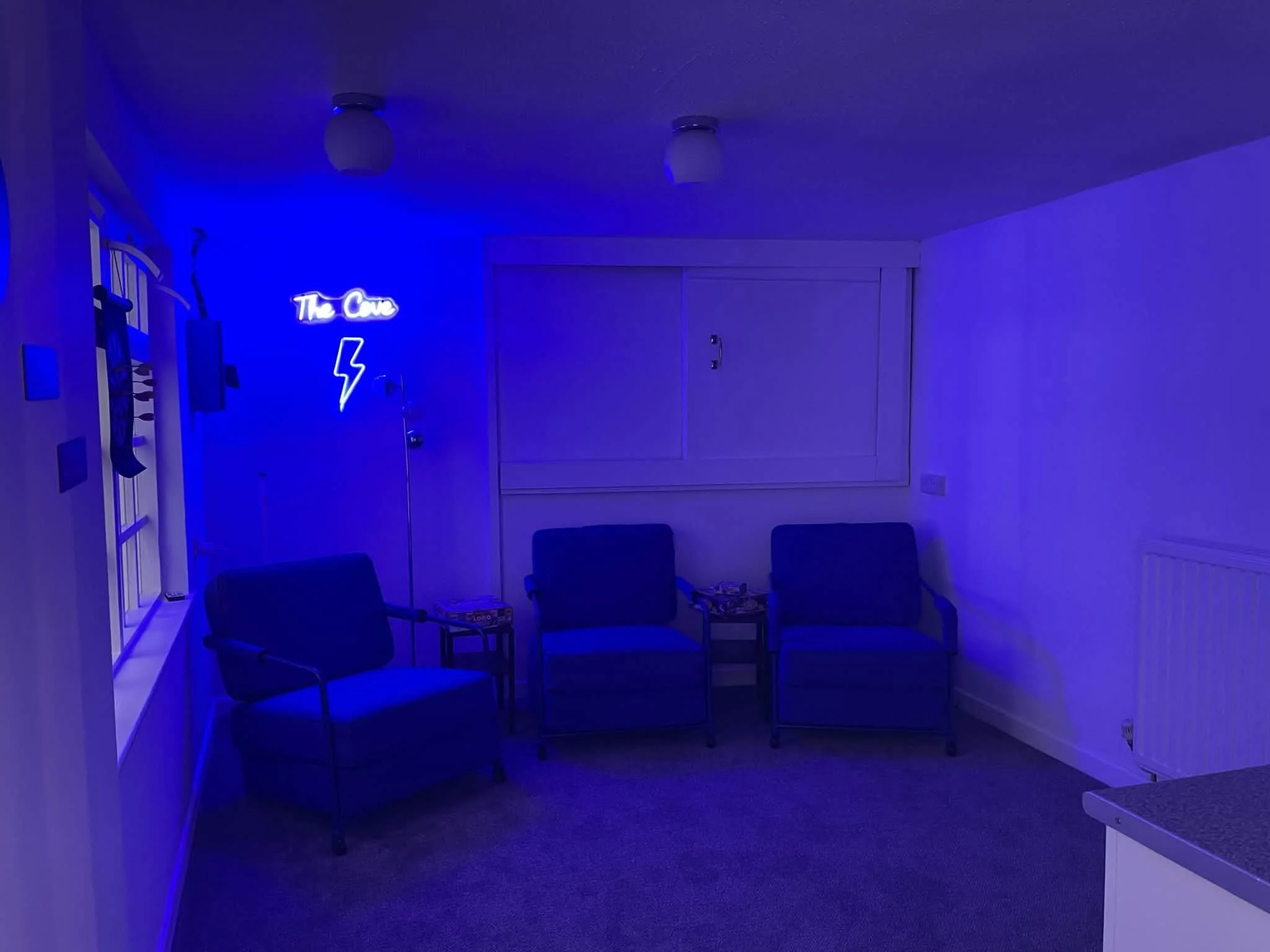 A room with blue lighting, four chairs arranged in a semi-circle, a neon sign saying 'The Cave' with a lightning bolt, and a few boxes on the floor.