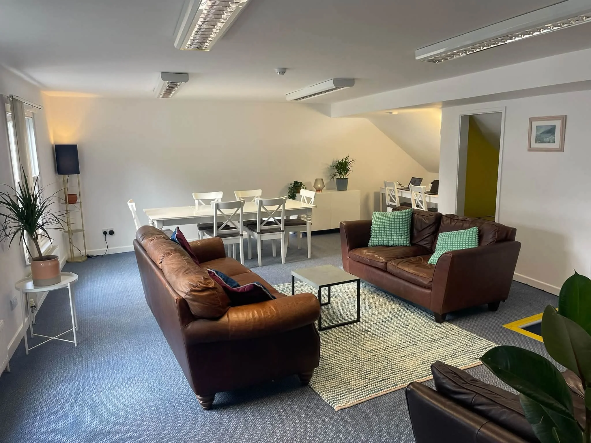 A cozy office or lounge area with two brown leather sofas, a white dining table with six chairs, and various indoor plants. The room has light-colored walls, a picture frame on the wall, and multiple ceiling lights.
