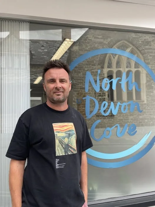 Man standing outside North Devon Cove window, wearing a black t-shirt with a colorful landscape print, in front of a window with the logo of North Devon Cove. Behind him, a stone building with a Gothic-style arched window.