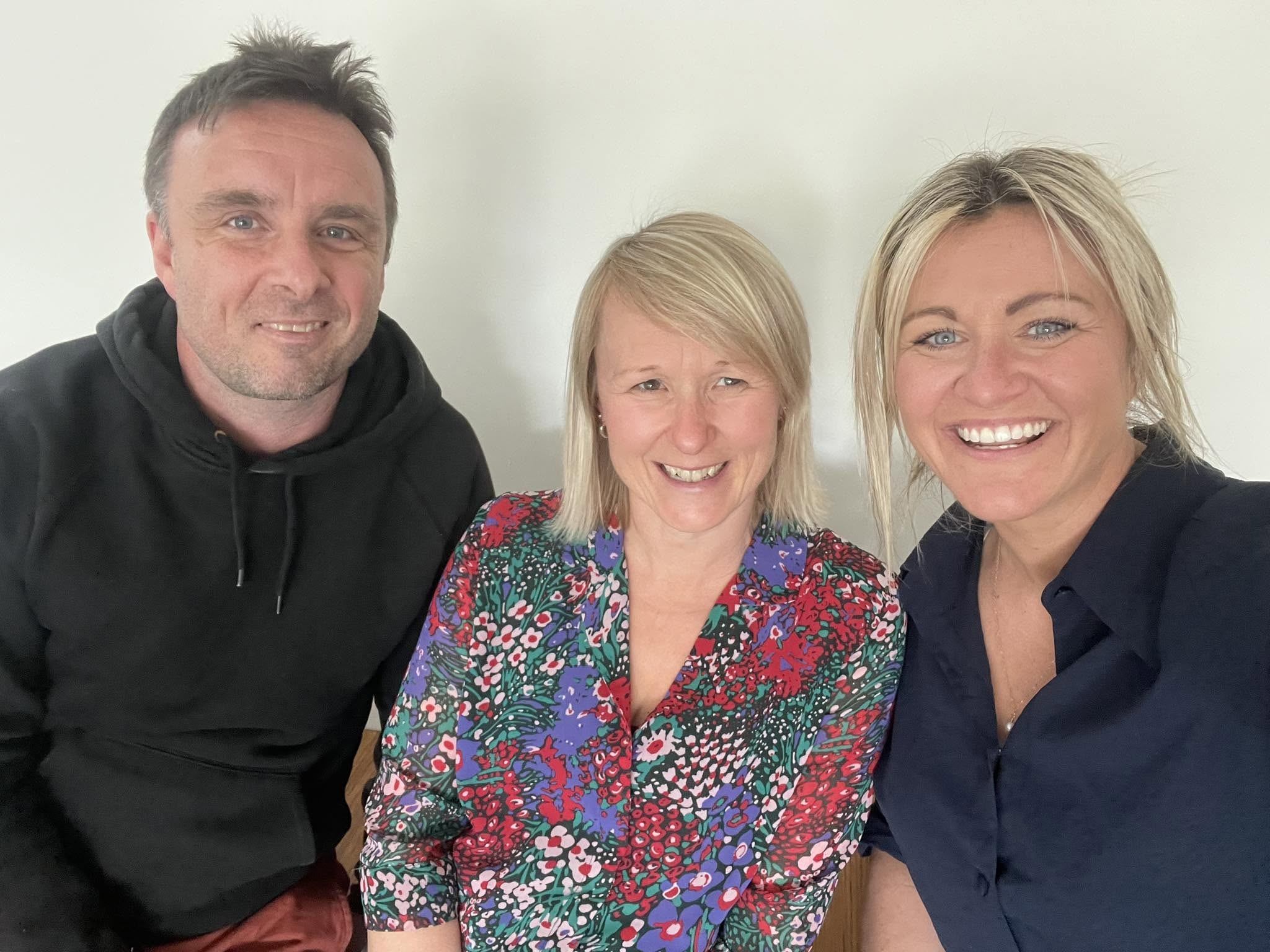 Three people, two women and one man, smiling and taking a selfie together against a plain white wall.