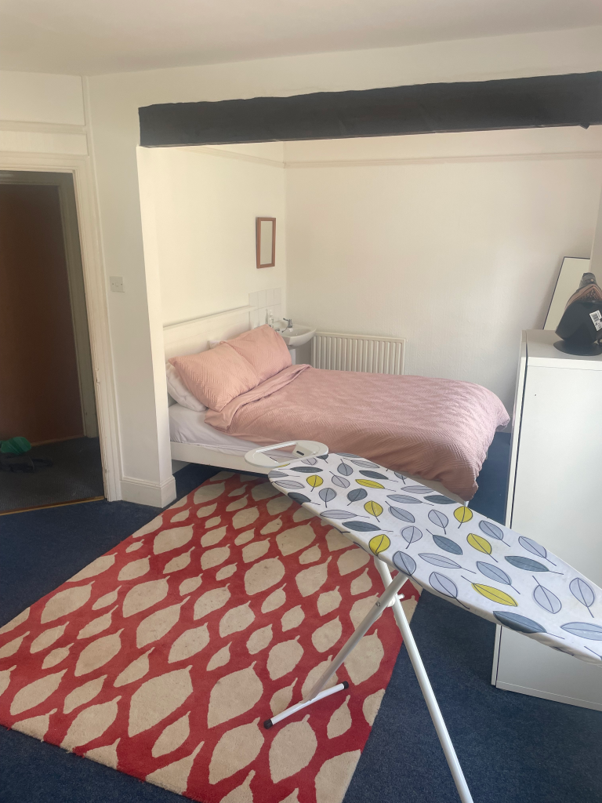 A bedroom with a bed in pink bedding, an ironing board with a patterned cover, and a red and beige patterned rug. There is a door to the left and a white dresser with a bag on top.