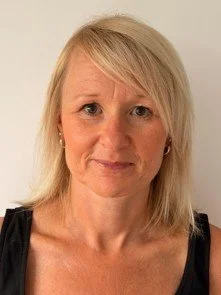 A woman with blonde hair and fair skin looking at the camera, wearing a black sleeveless top, standing against a white background.