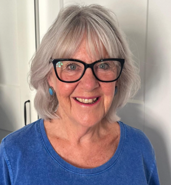 An elderly woman with gray hair, glasses, and blue earrings smiling at the camera, wearing a blue top.