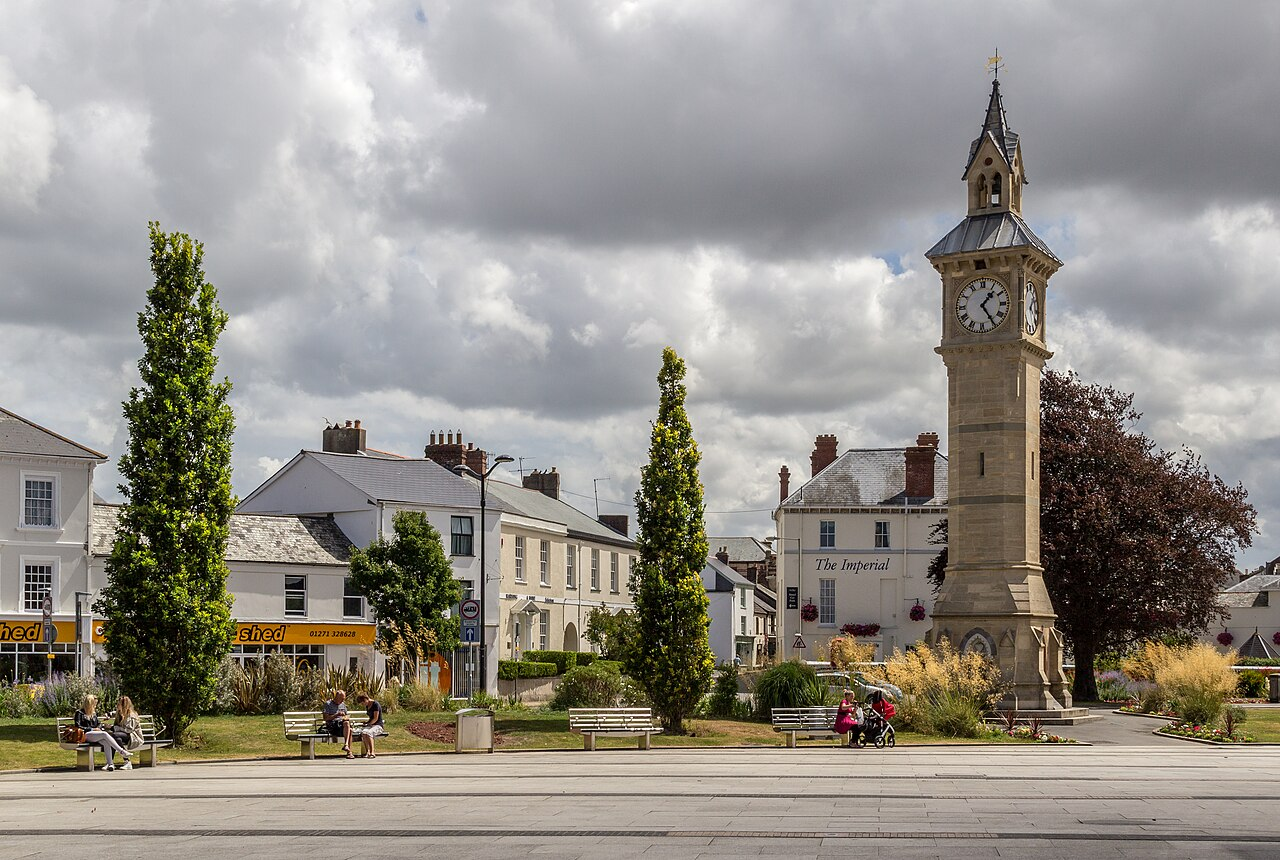 A public square with a clock tower, benches, trees, and people sitting and walking, under a cloudy sky.