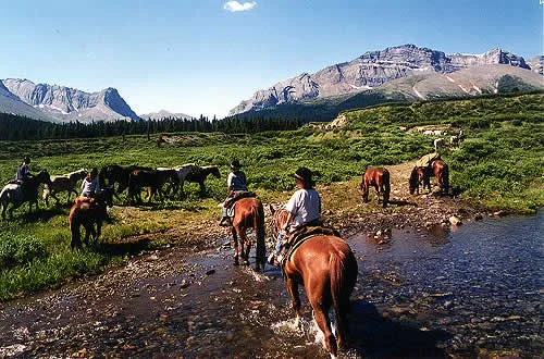 Horseback riders crossing a shallow stream in a mountain valley beneath rugged alpine peaks